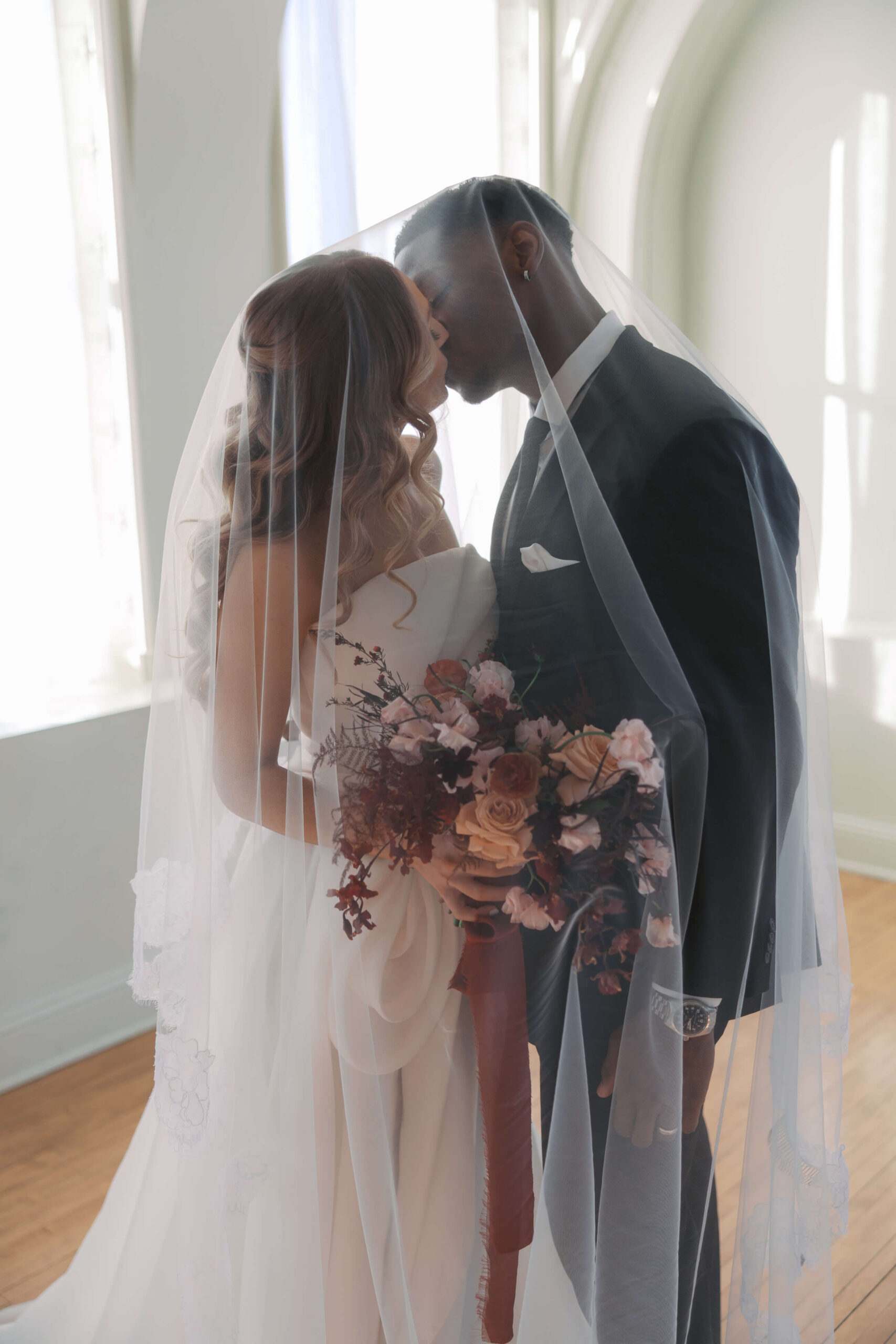 A bride and groom kiss while hiding under the veil in a ballroom