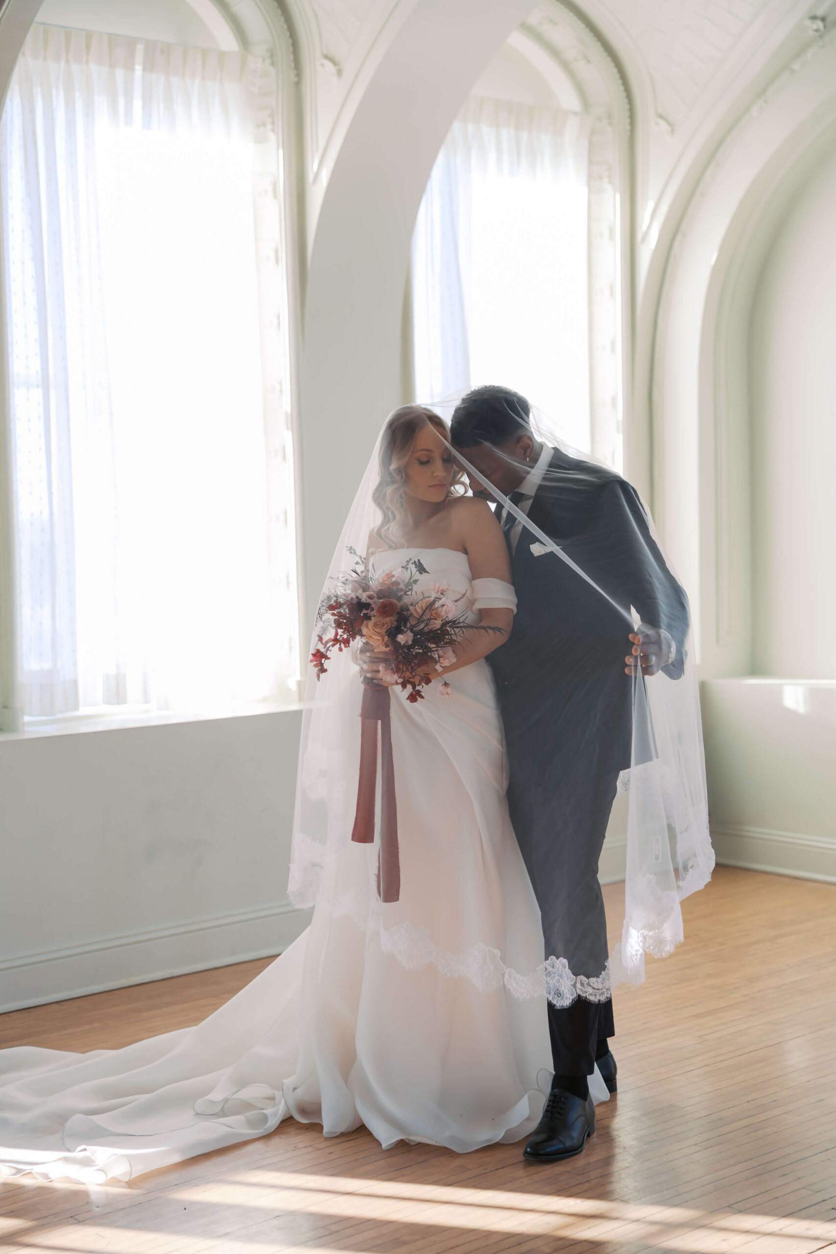 A bride and groom snuggle under the long veil under a window while holding a bouquet at their cottonwood hotel wedding