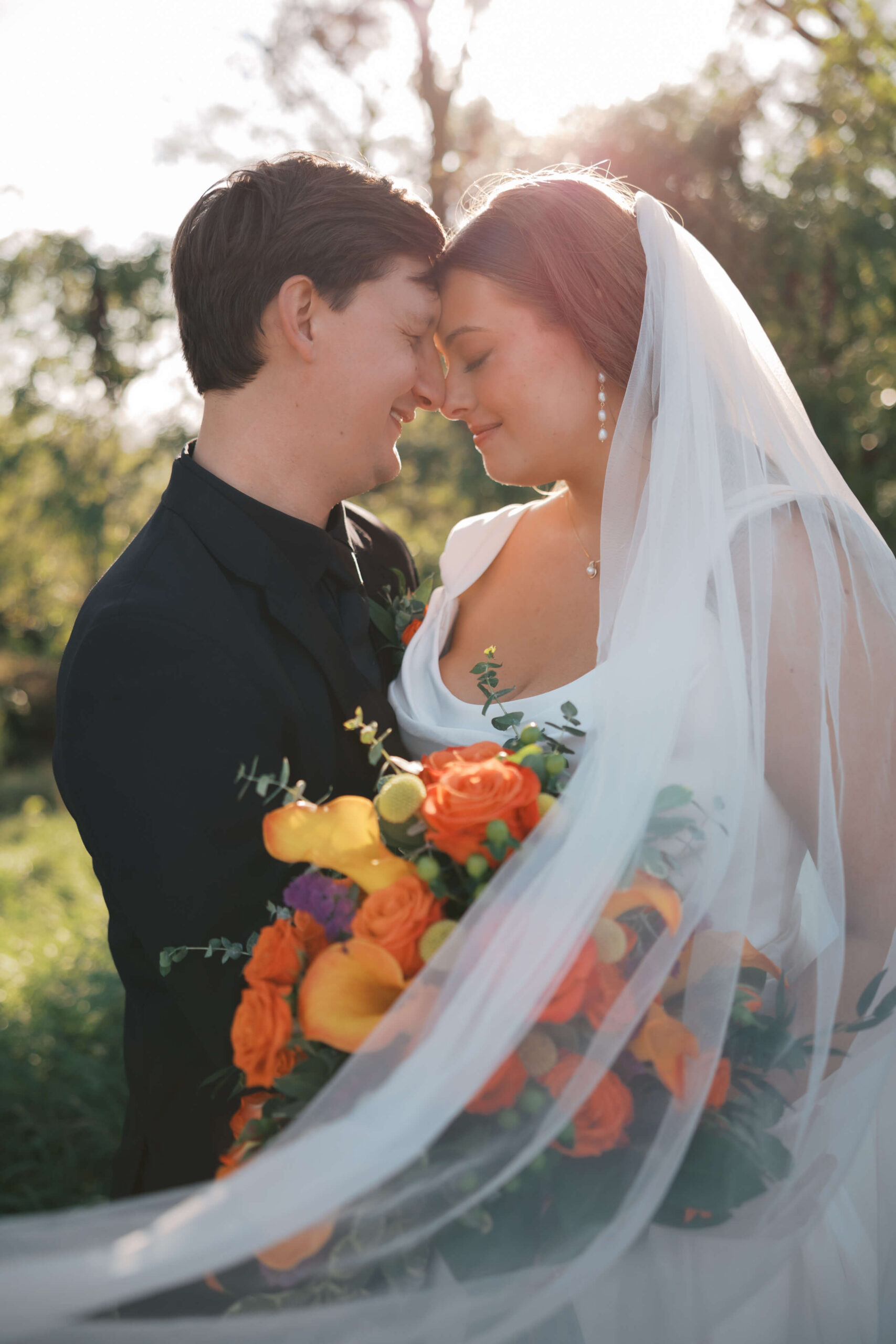 Newlyweds nuzzle at sunset while holding a colorful bouquet as the veil wraps around them during their glacial till vineyard wedding