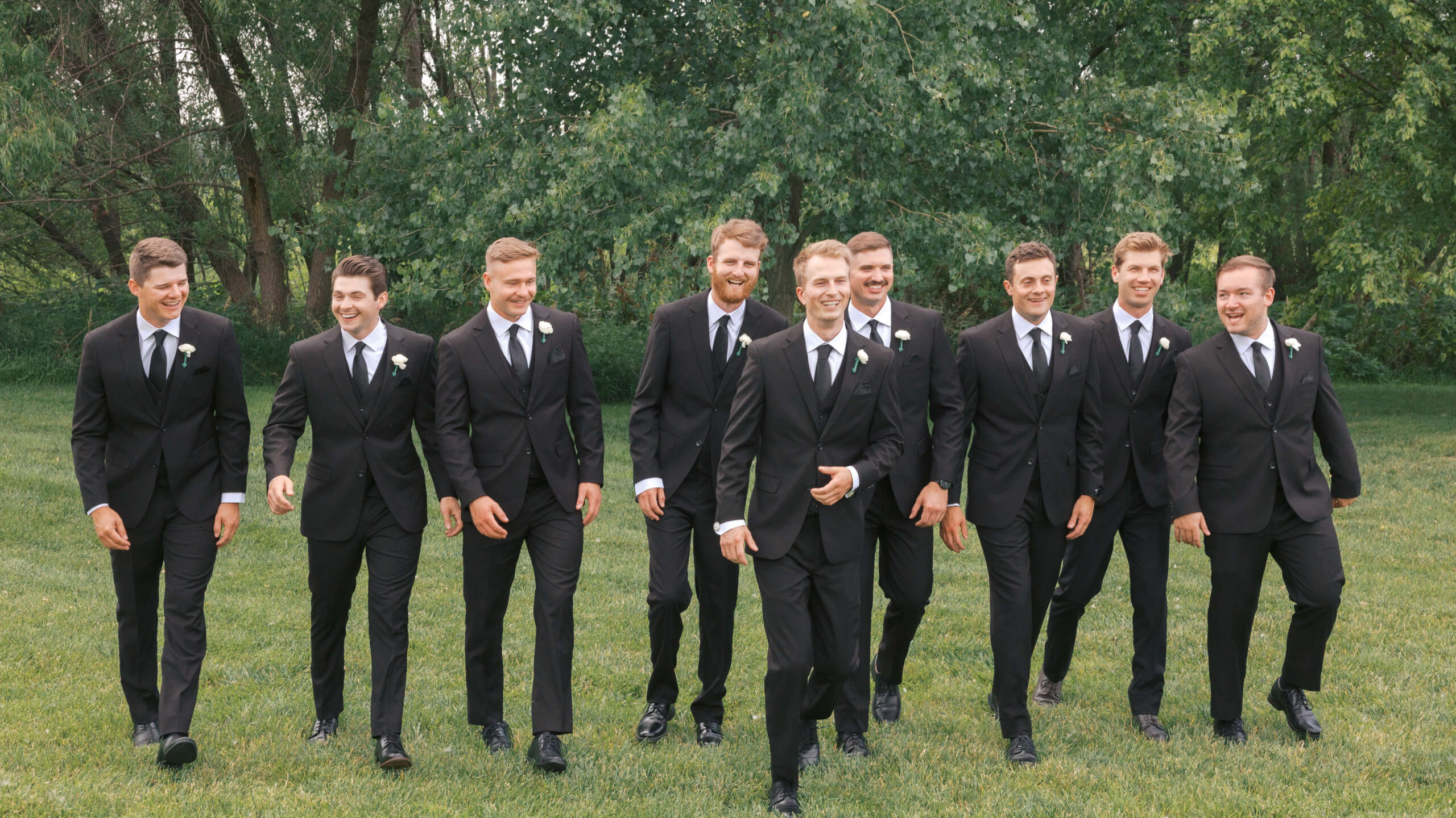 A groom in black walks in front of his groomsman in matching suits in a lawn at sunset