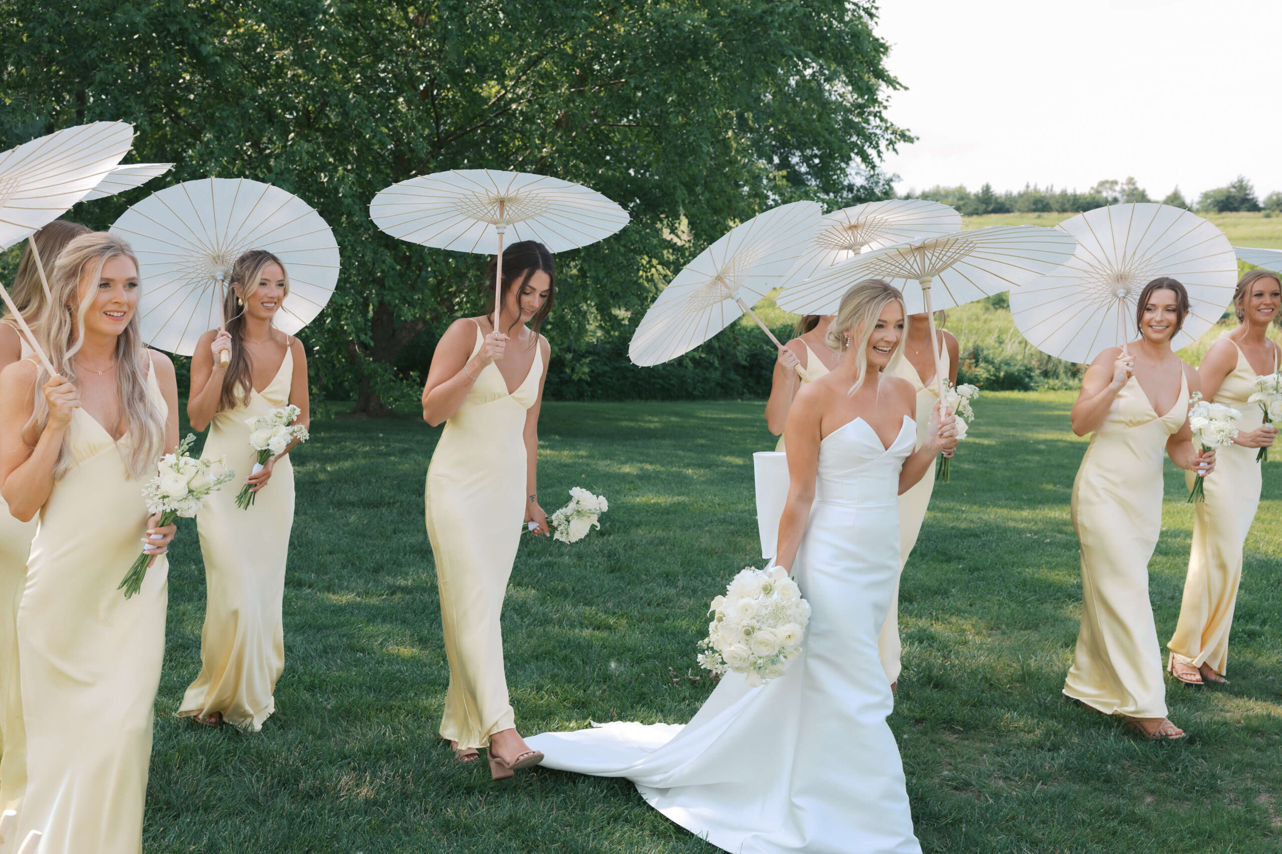 A happy bride walks with her bridesmaids all in yellow holding white parasols in a lawn during her happy hollow club omaha wedding