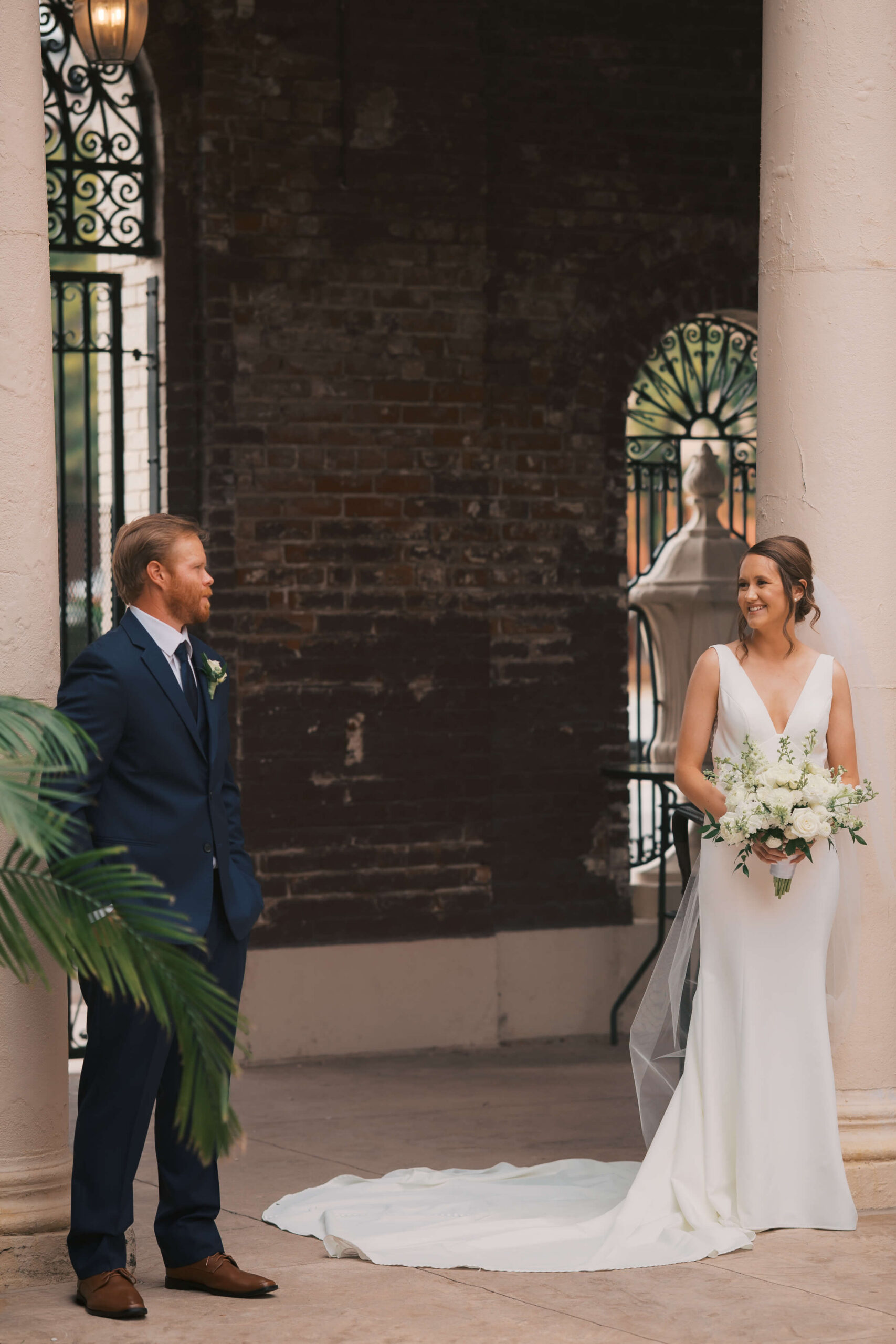 A bride smiles big while standing and admiring her groom on a patio between columns