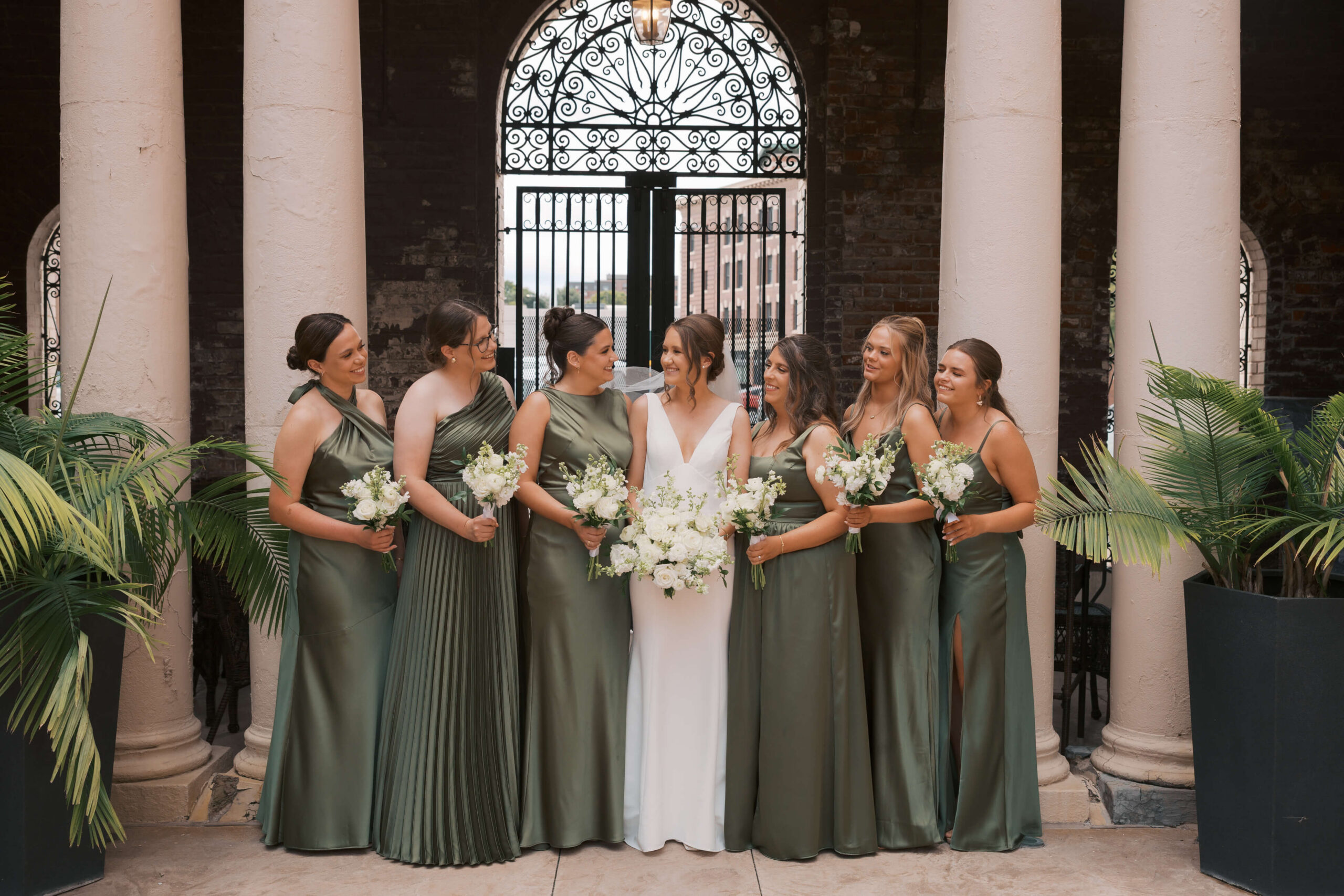 A happy bride laughs with her bridesmaids standing between columns of the magnolia hotel wedding venue holding their bouquets in green gowns