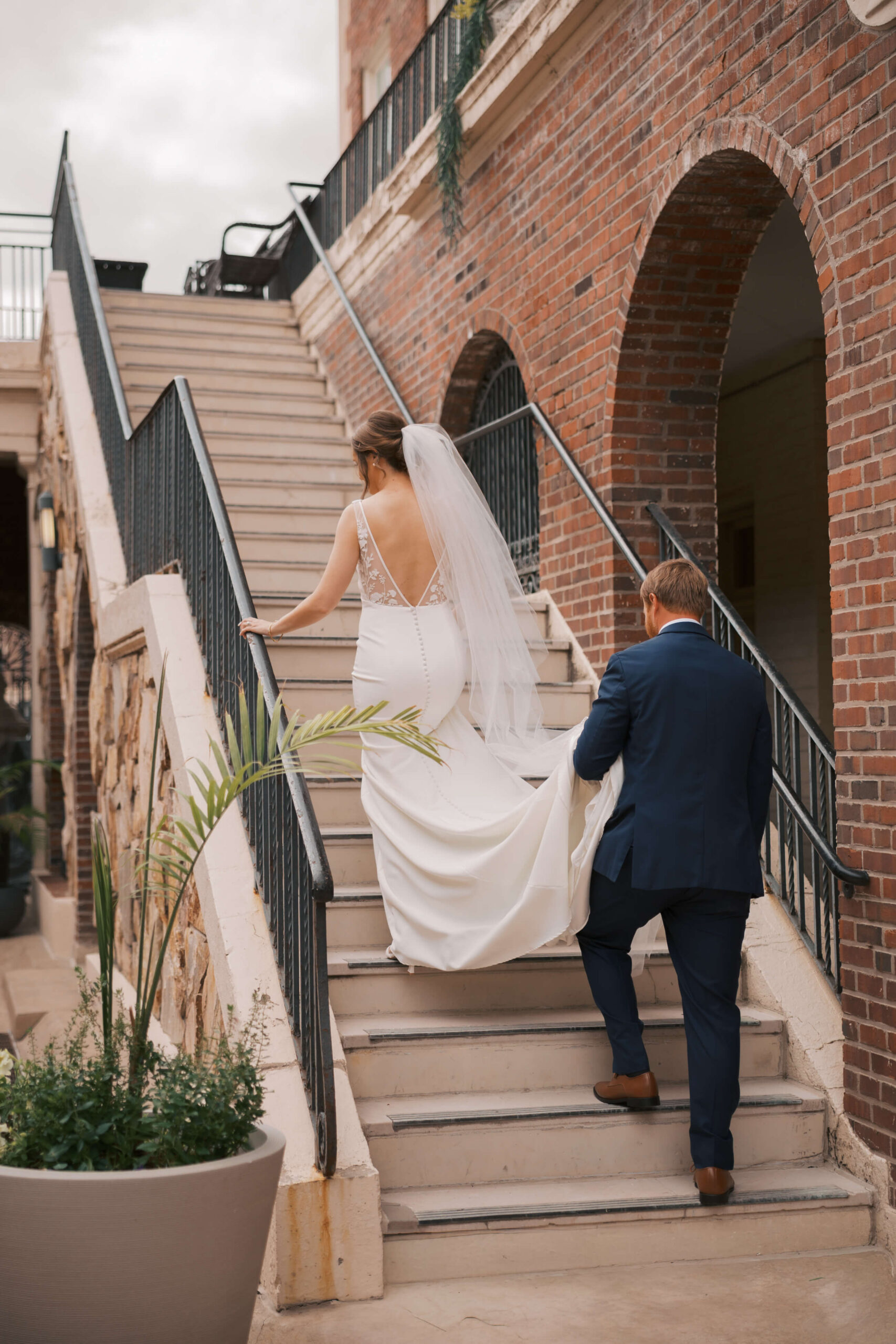 A groom holds his bride's train as they walk up stairs in the patio of the magnolia hotel wedding venue