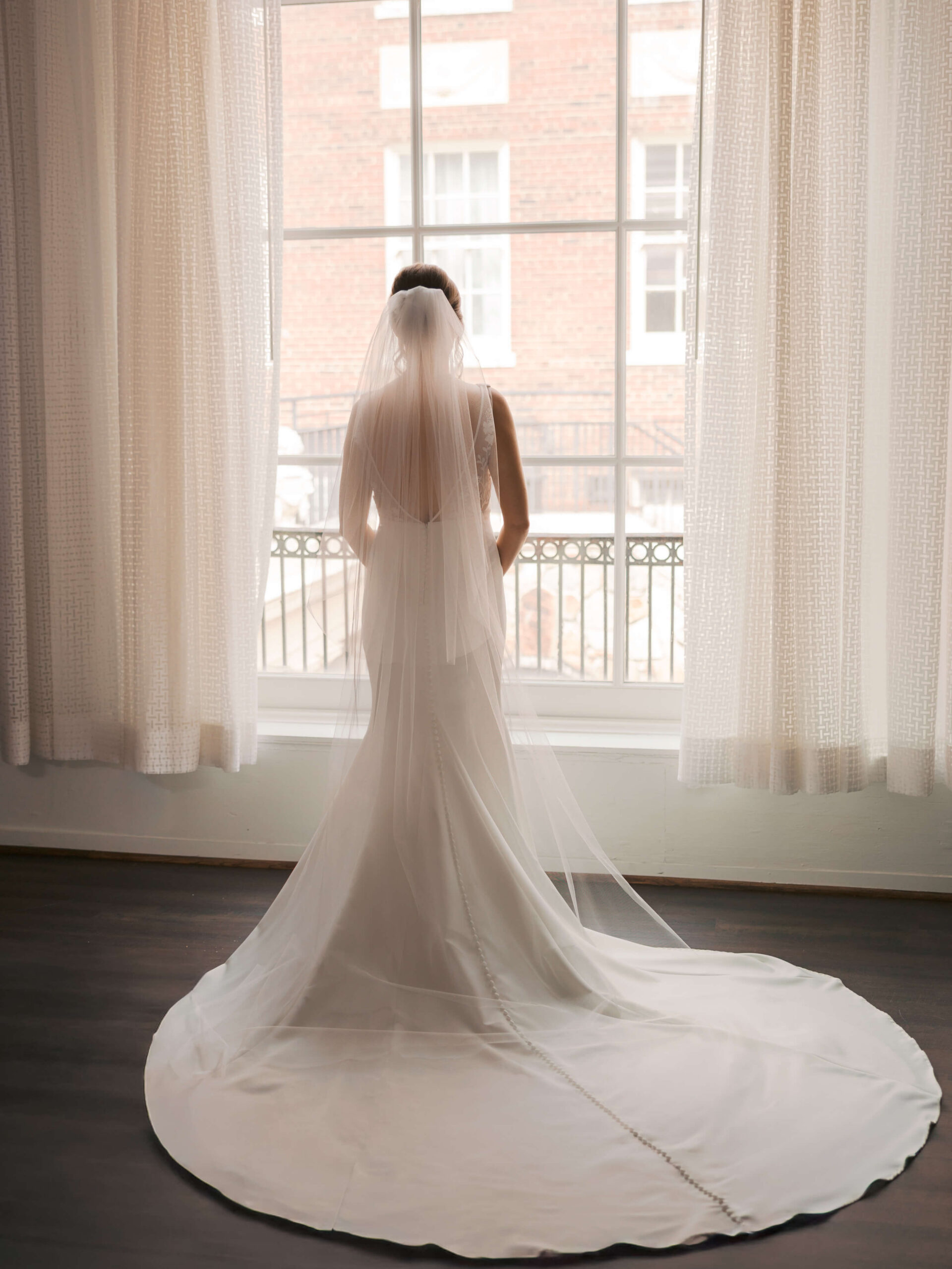 A bride stands in a tall window gazing out with train spread at the magnolia hotel wedding venue