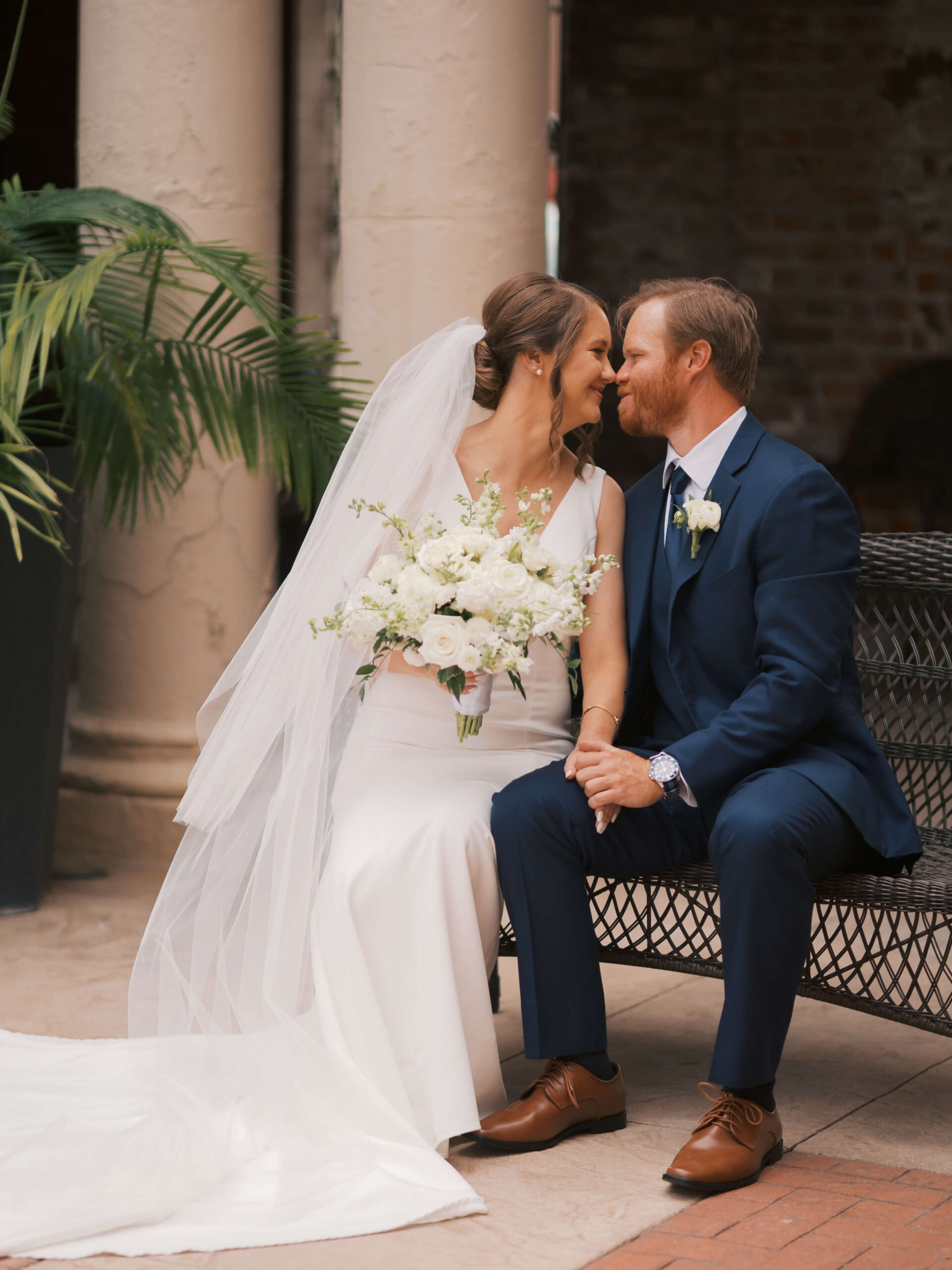 A bride and groom sit on a metal bench holding hands and leaning in for a kiss