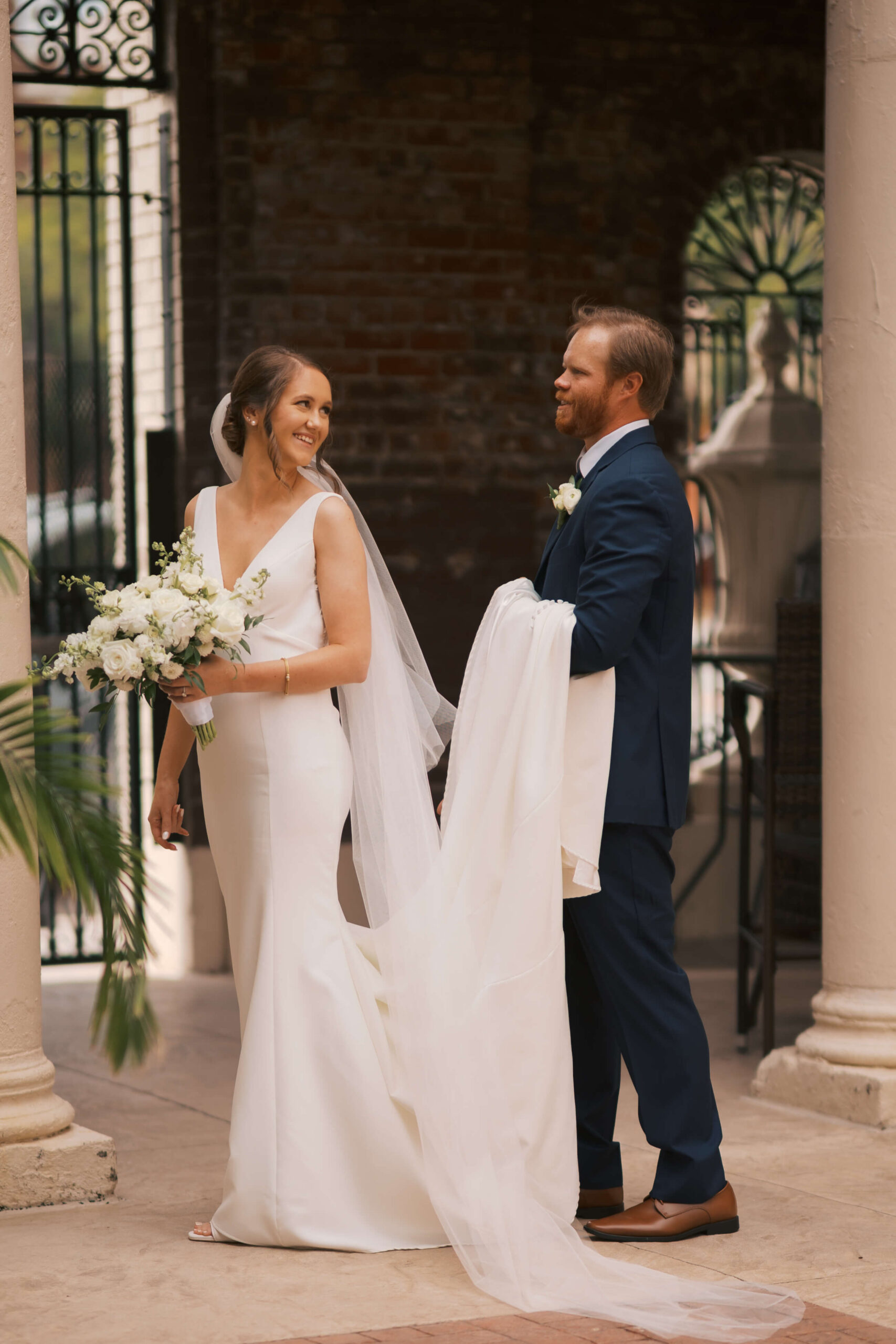A bride smniles over her shoulder to her groom holding her train as they walk together during their magnolia hotel wedding