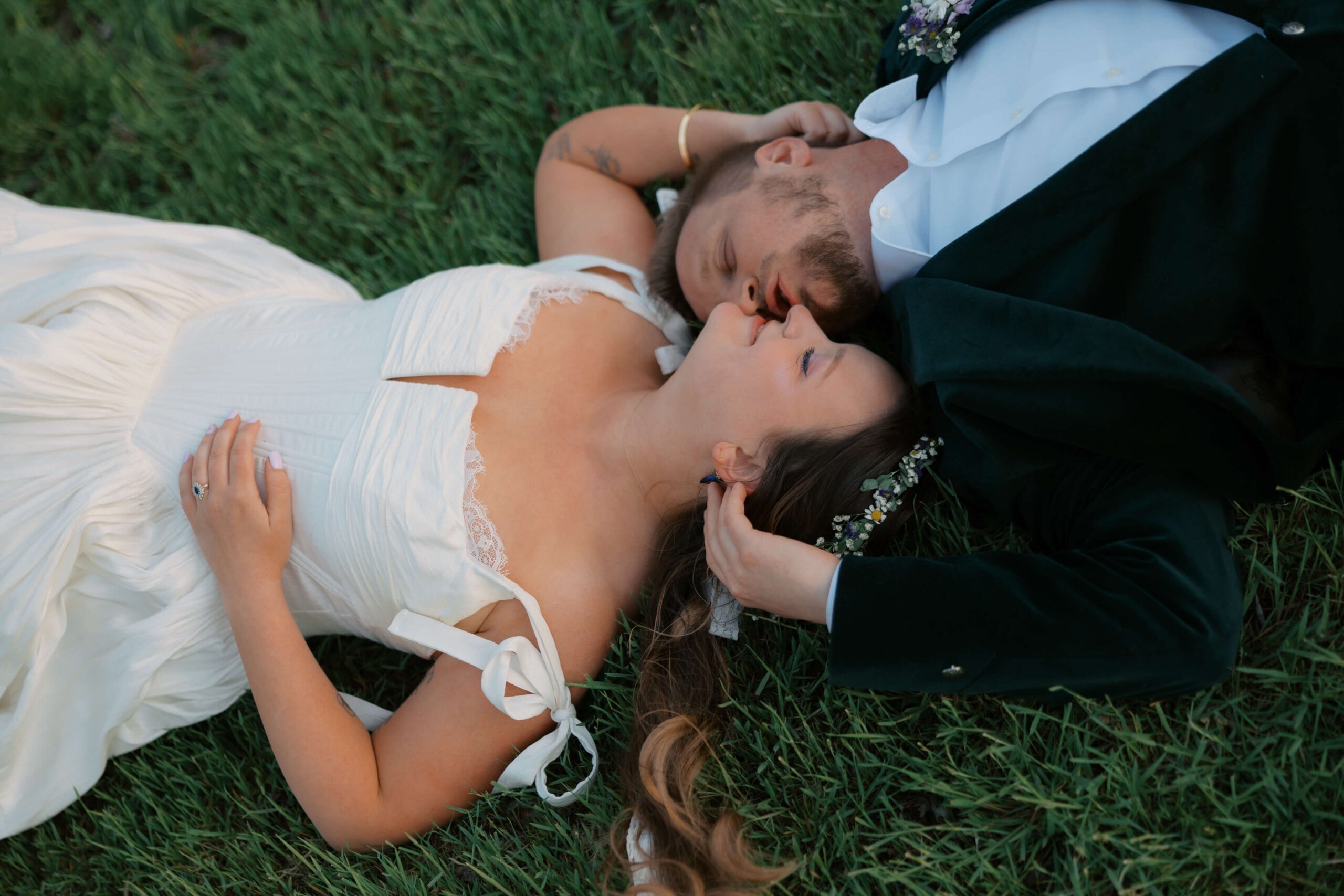 Newlyweds lean in for a kiss while laying head to head in the grass on their backs