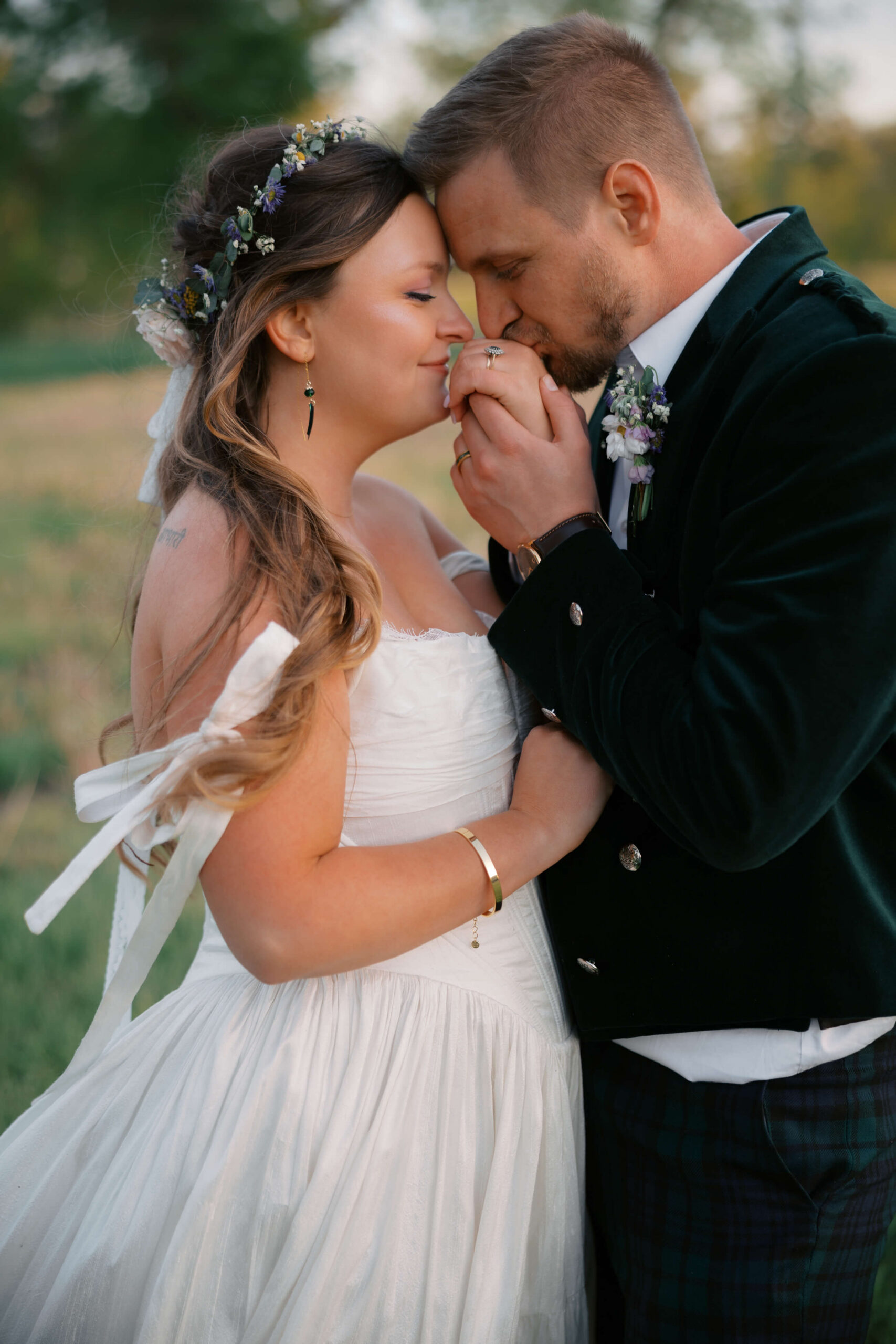 A happy groom kisses the hand of his bride and they stand close in a field at sunset at the northern lights venue