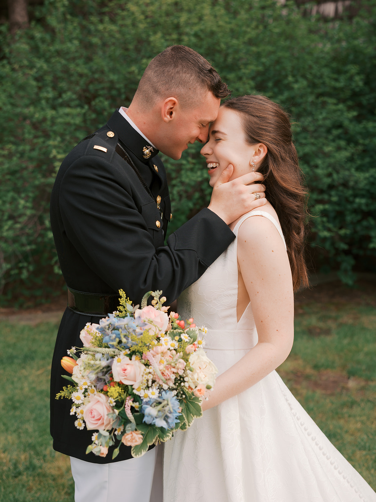 Happy newlyweds giggle and laugh while leaning in for a kiss in a lawn holding a bouquet during their Quarry Oaks wedding