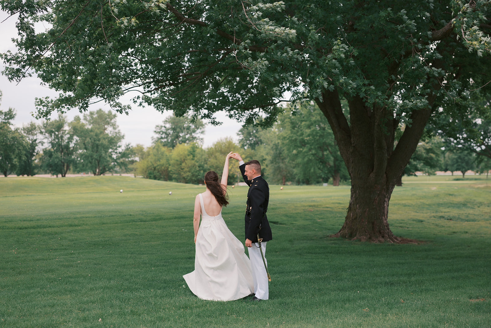 A groom in his Marines formal uniform dances with his bride under a tree on a golf course at their Quarry Oaks wedding