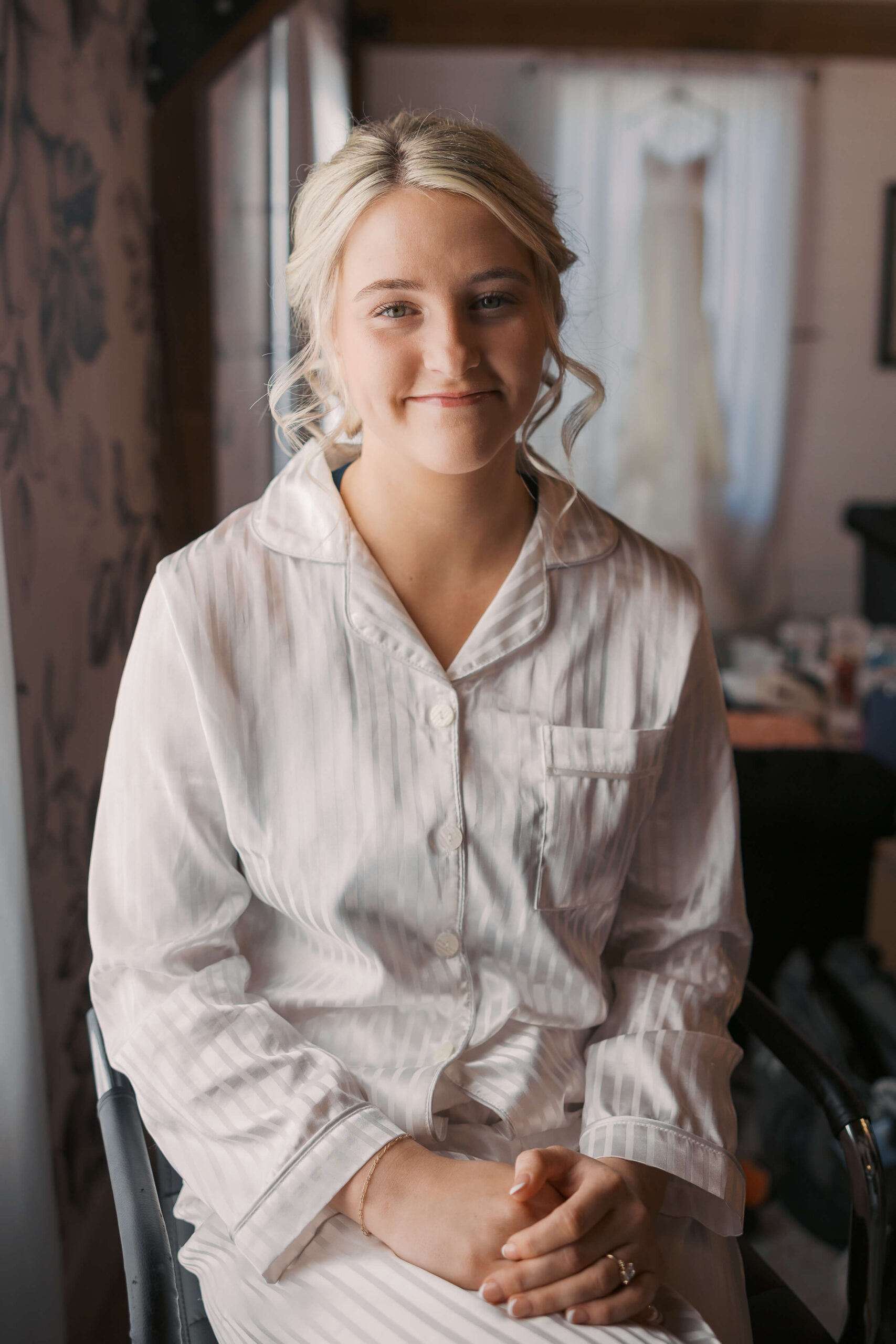 A bride smiles while sitting in white pajamas in the getting ready suite with hands in her lap