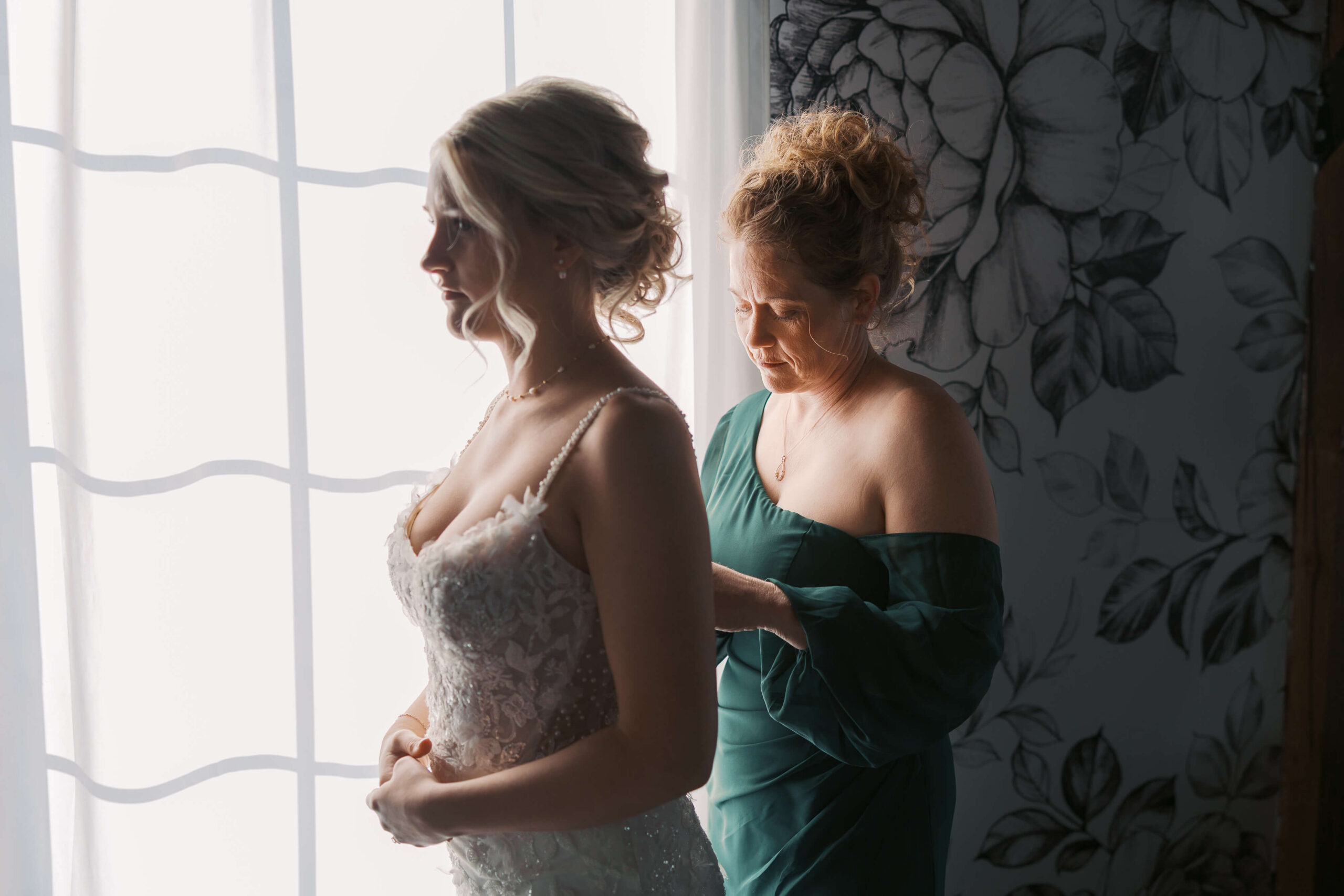 Mom in green helps the bride into her gown while standing in a window at Seven Willows wedding venue