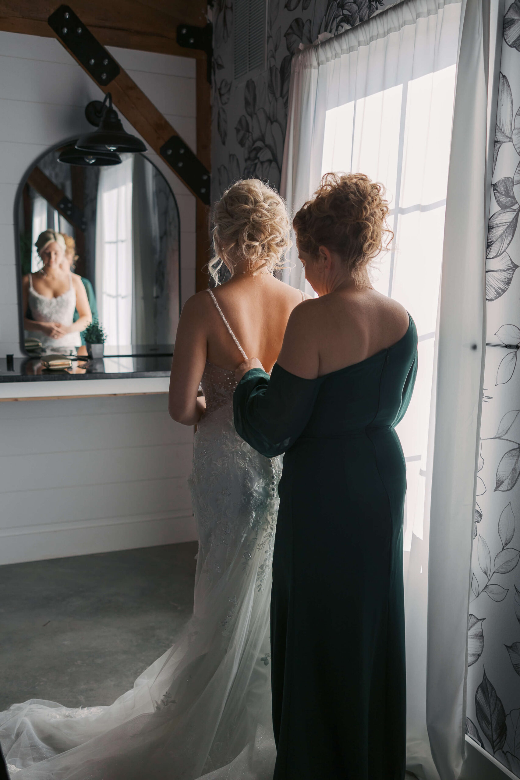 Mom helps button in her daughter into her wedding gown in a mirror and window at Seven Willows wedding venue