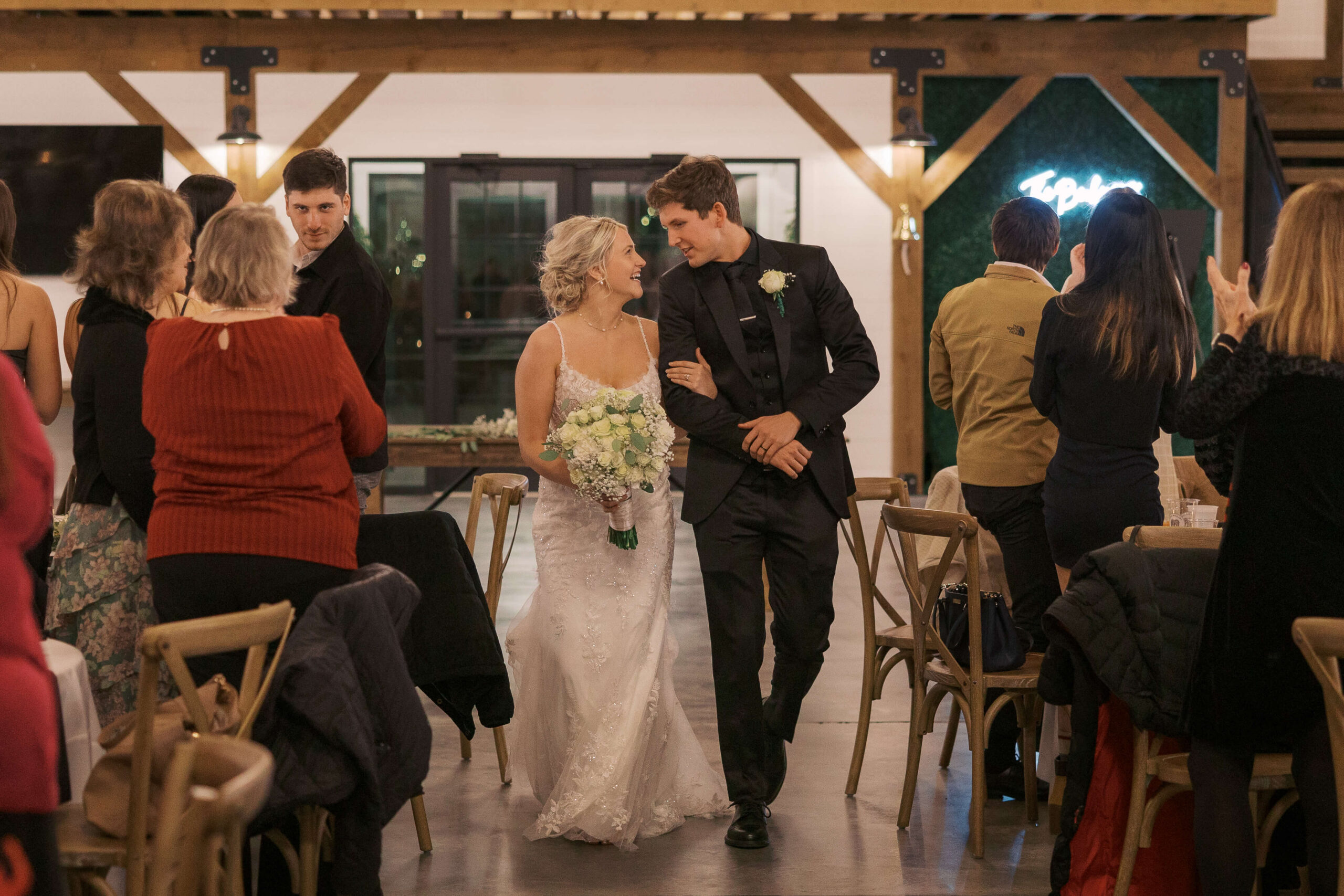 A bride and groom walk arm in arm to enter their wedding reception at Seven Willows wedding venue as guests stand and clap