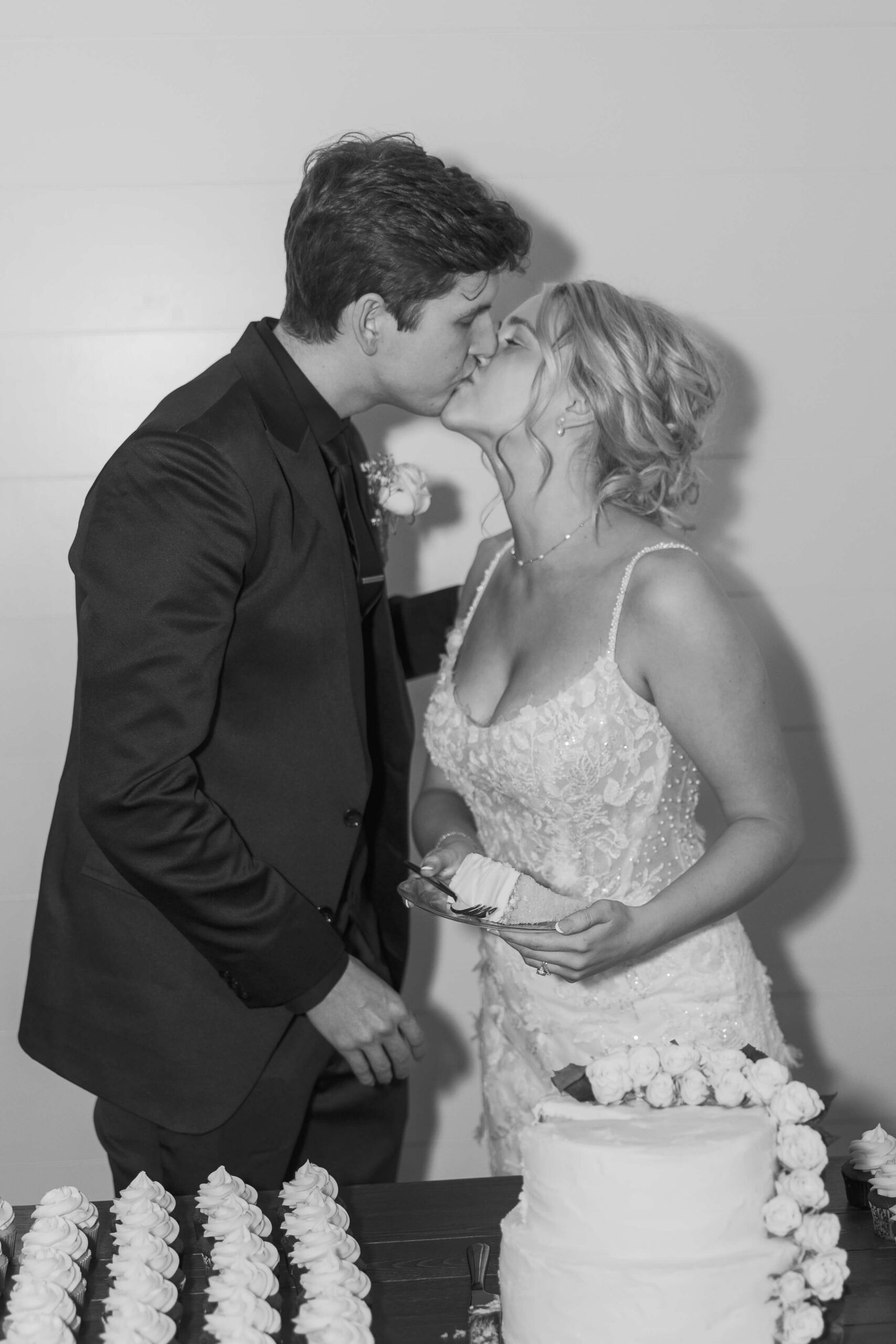A black and white image of a bride and groom kissing behind their cake table