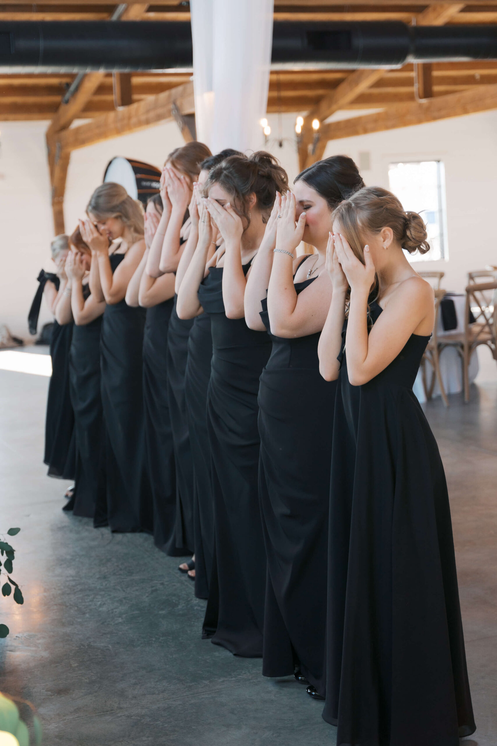 Bridesmaids in black dresses cover their eyes for their first look with the bride in a reception space