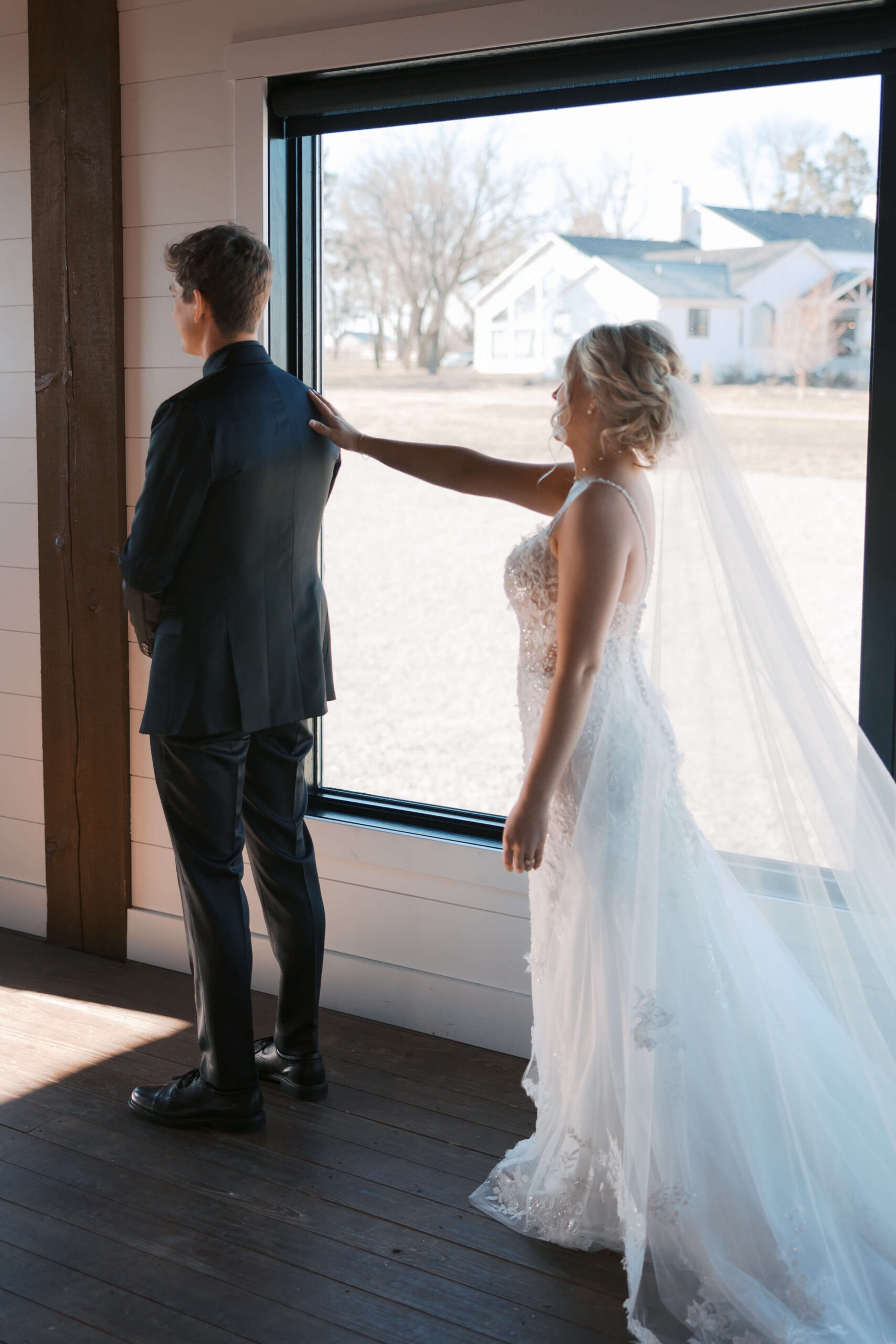 A bride reaches out for a first look in a window touching her groom's back shoulder