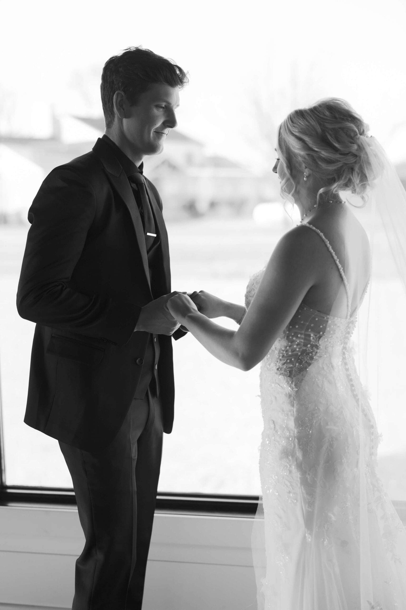 A black and white image of a bride and groom holding hands in a large window smiling at each other at the Seven Willows wedding venue