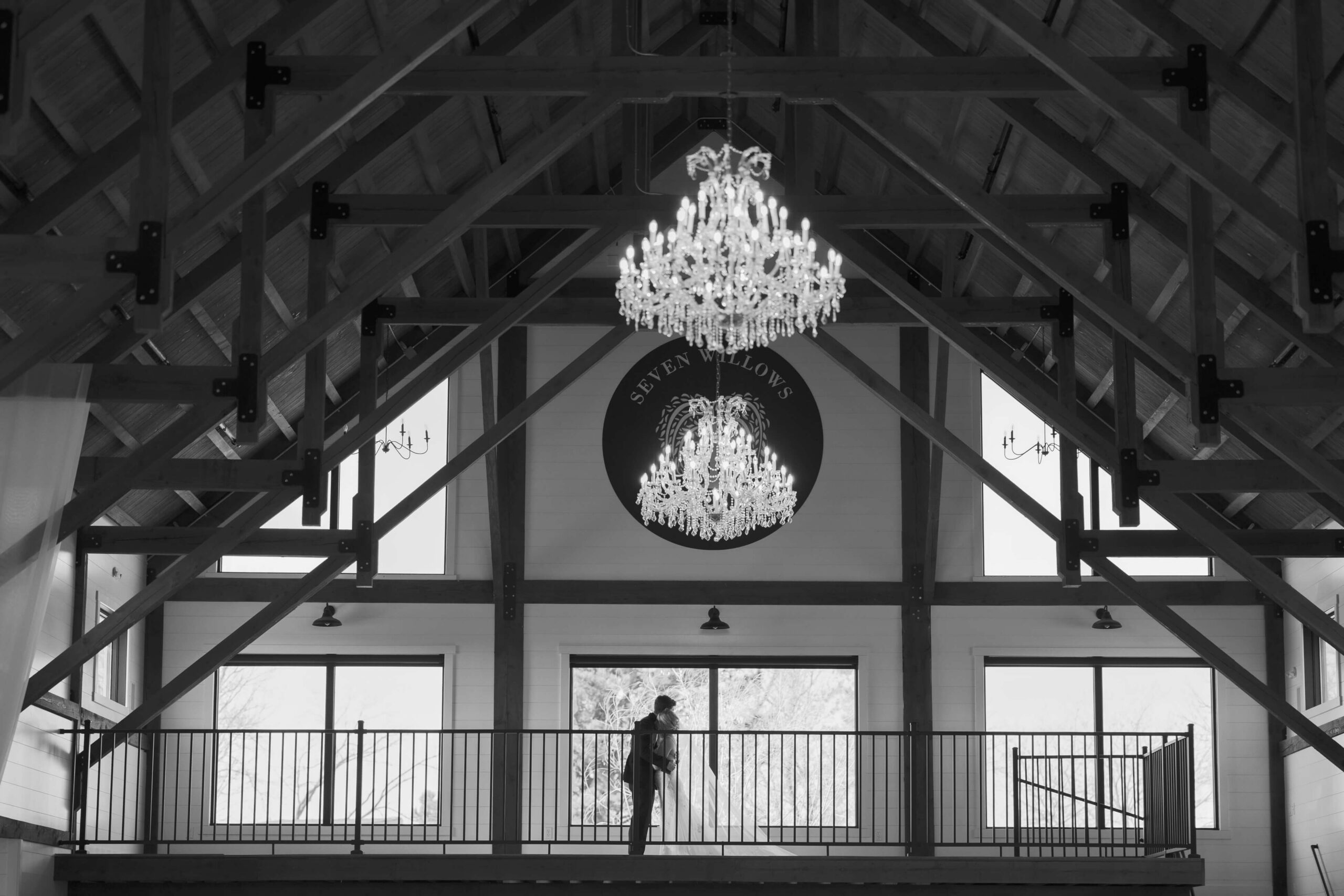 Newlyweds embrace in black and white on a balcony under crystal chandeliers at the Seven Willows wedding venue
