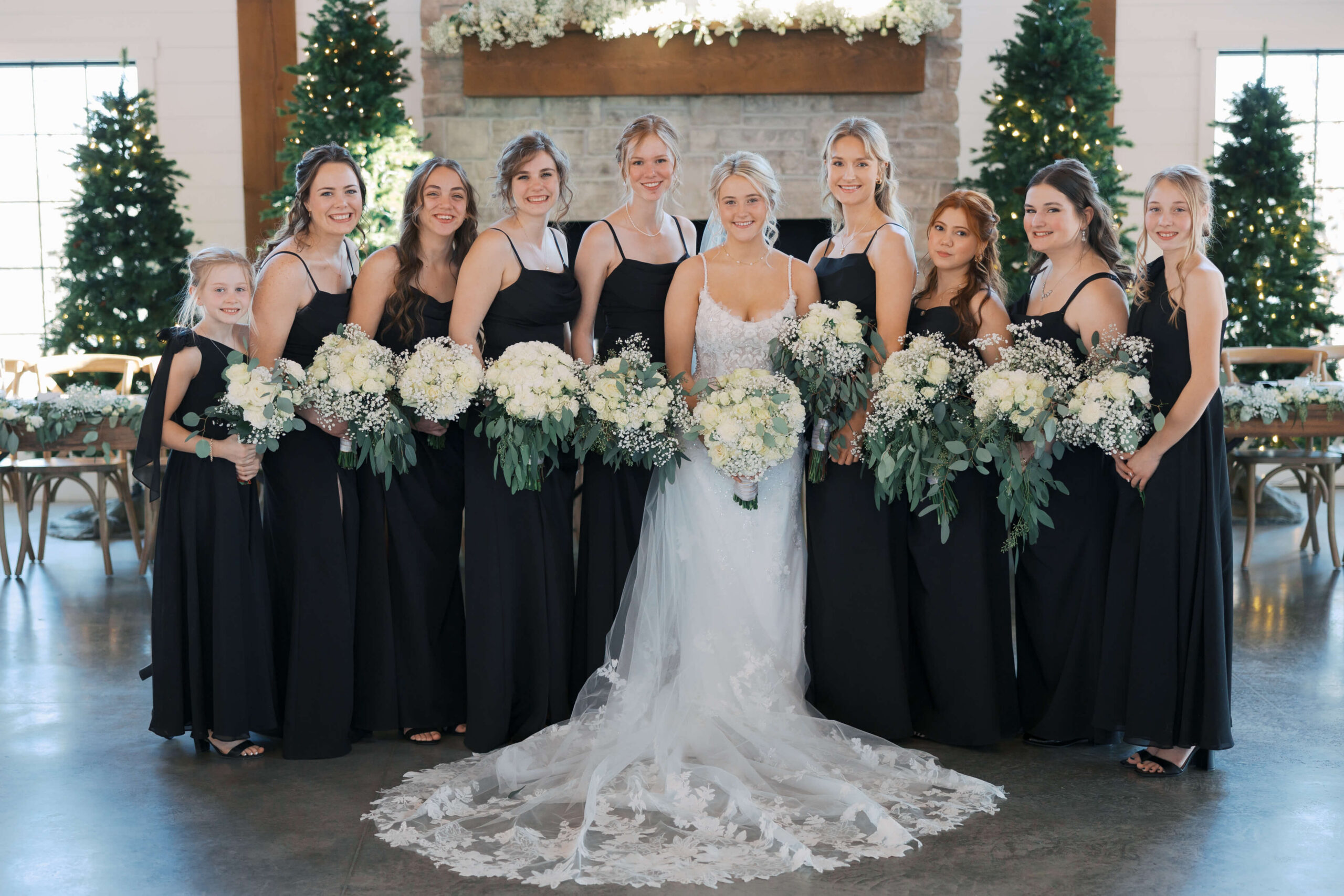A smiling bride in a lace gown stands with her bridesmaids in black holding matching white rose bouquets in a reception space