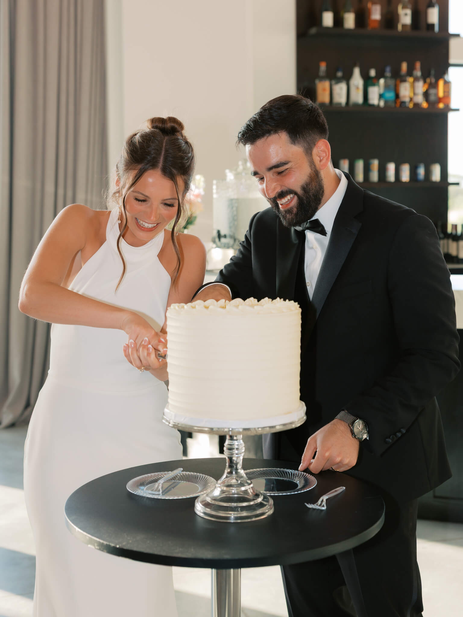 A bride and groom laugh while cutting their tall one tier cake