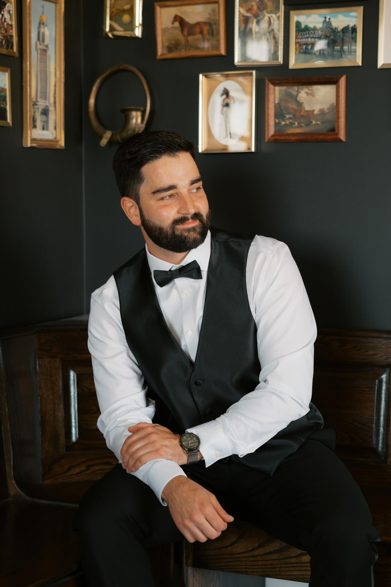 A groom sits on a wooden bench in his tuxedo vest while getting ready