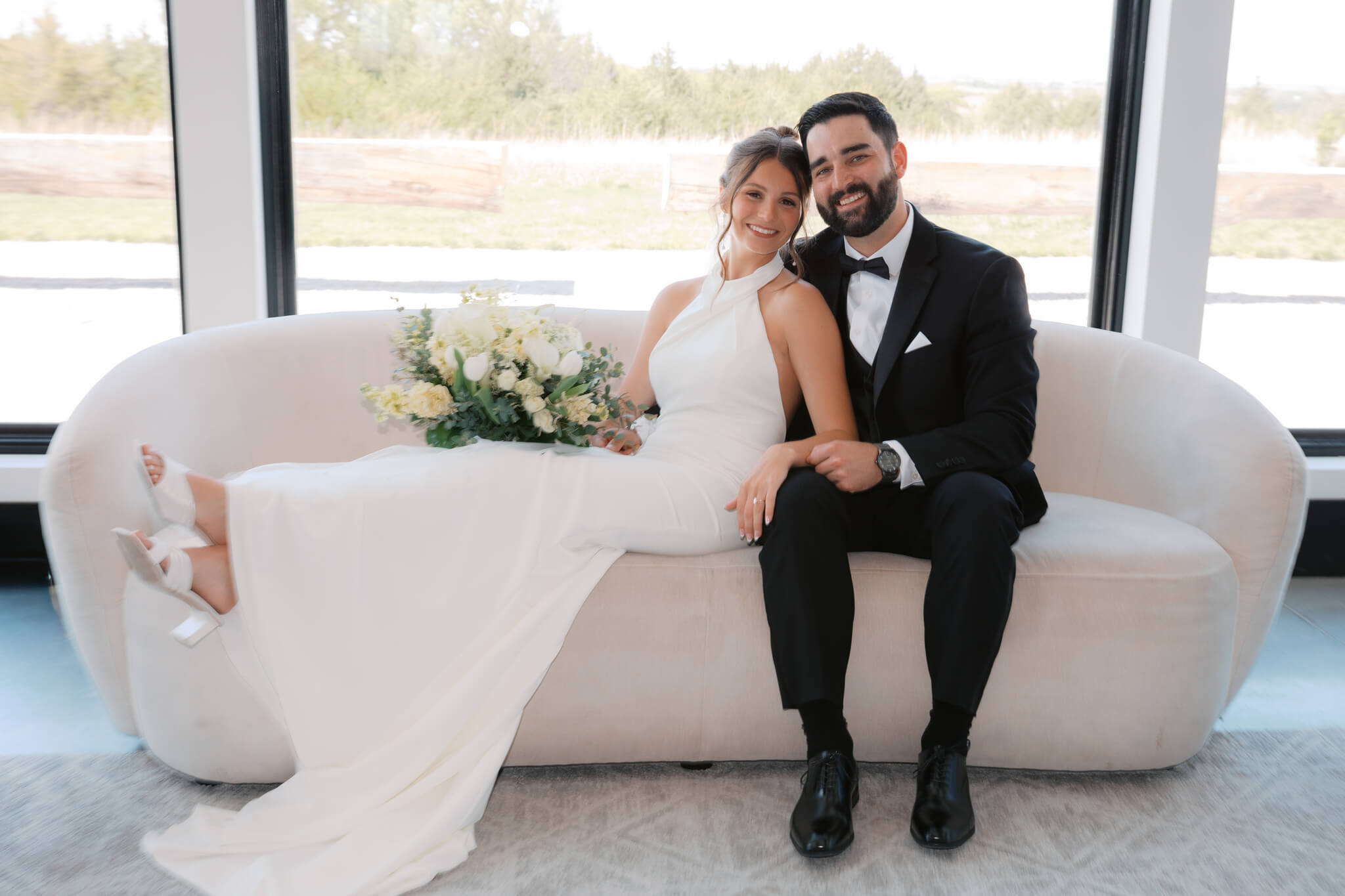 A bride lounges across a couch leaning against her groom in a large window