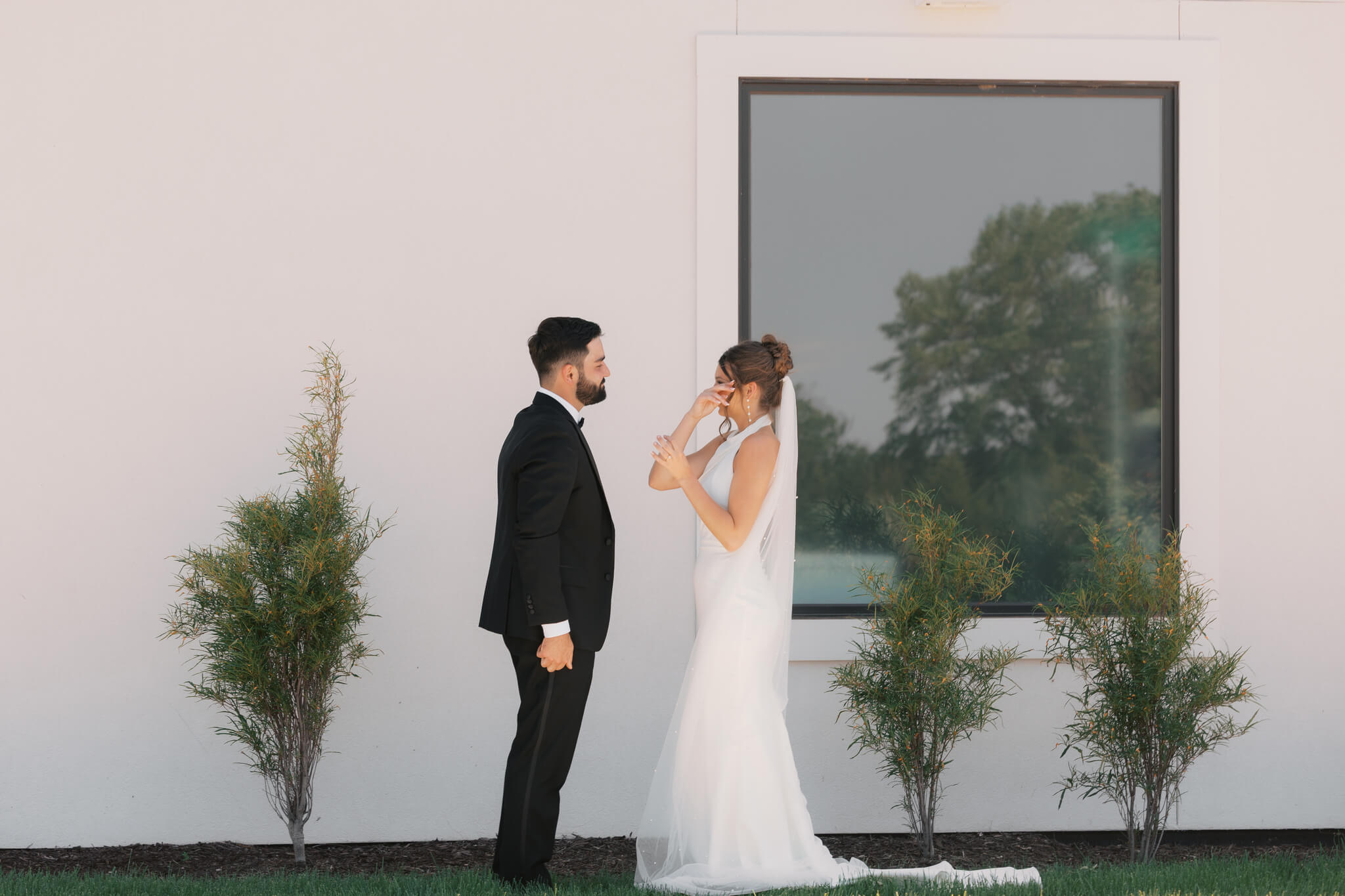 A bride wipes away tears while enjoying her first look at her groom in his black tux in a garden