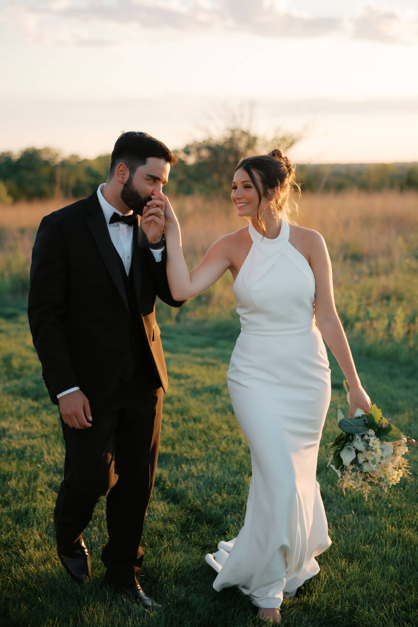 A groom in a black tux kisses the hand of his bride as they walk at sunset in the grass of The Acreage wedding venue