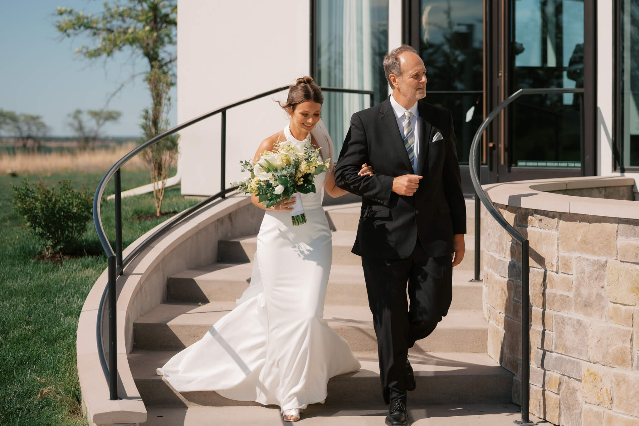A dad in a black suit walks his daughter down the stairs to her outdoor ceremony at The Acreage wedding venue