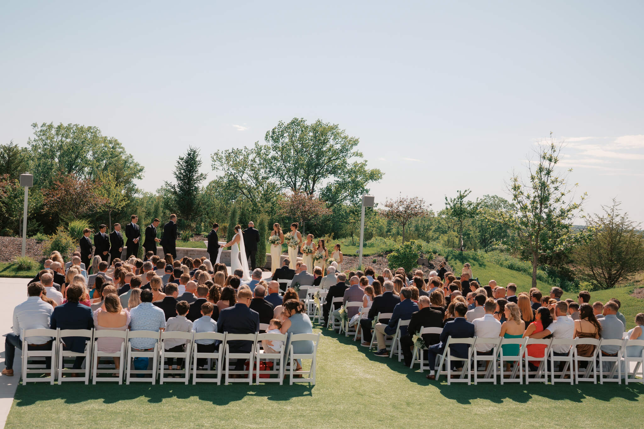 A wide look at a beautiful outdoor ceremony in the gardens of The Acreage wedding venue as the bride and groom hold hands