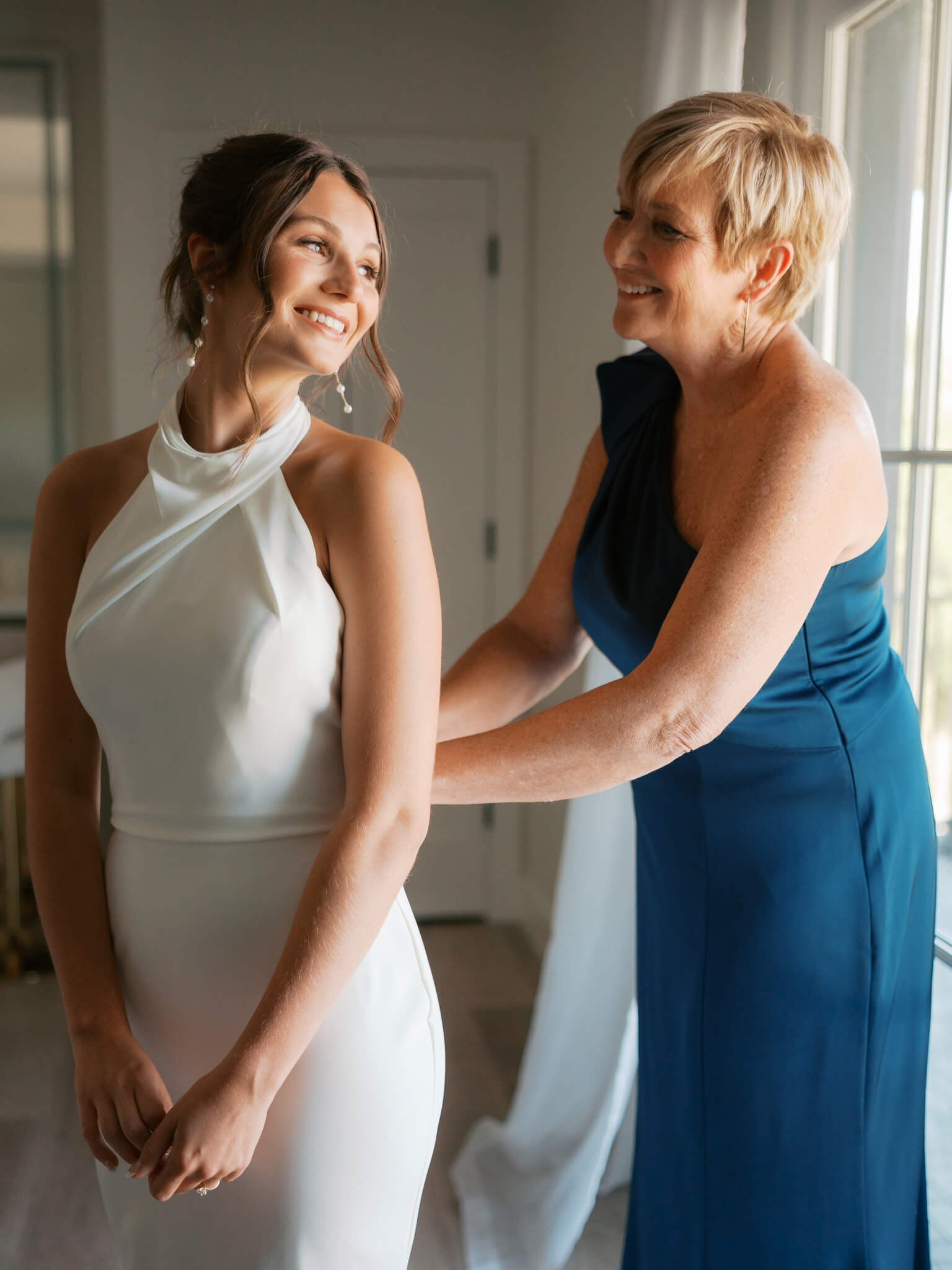 A bride smiles over her shoulder to mom helping with her dress in the getting ready suite