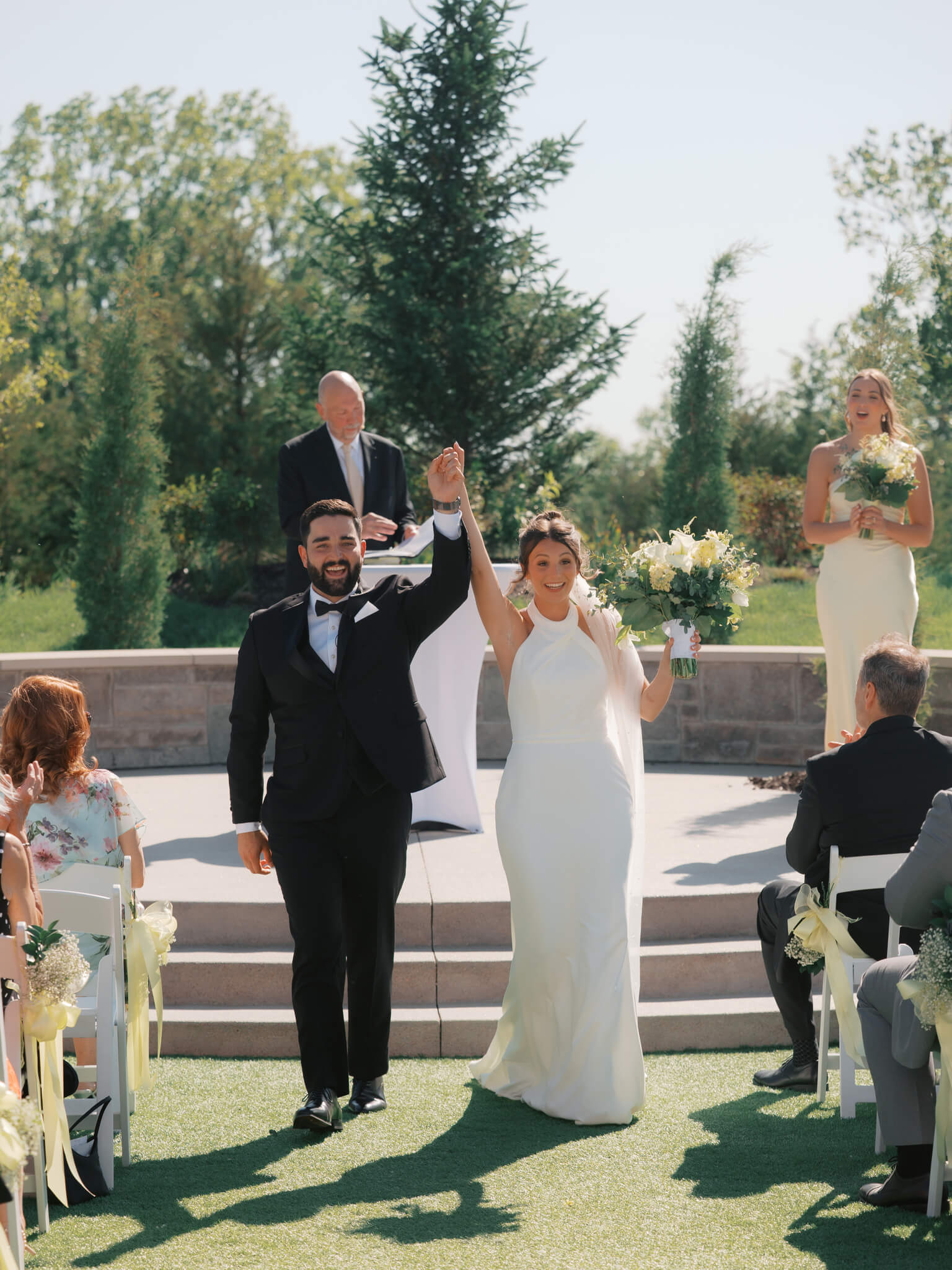 Newlyweds celebrate with arms raised while walking up the aisle of their outdoor ceremony