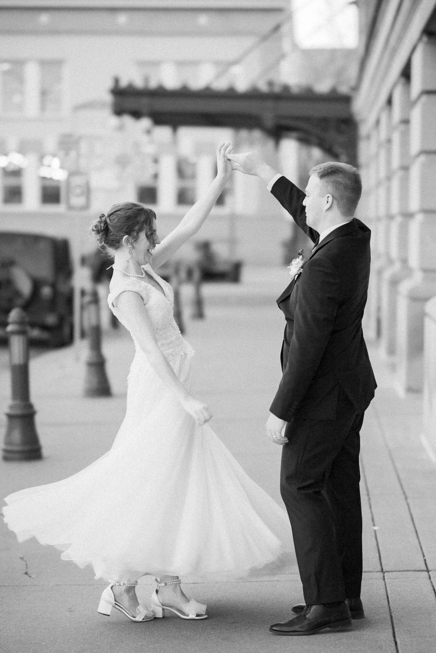 A bride and groom dance and twirl on the sidewalk while enjoying their empire room wedding in a black and white image
