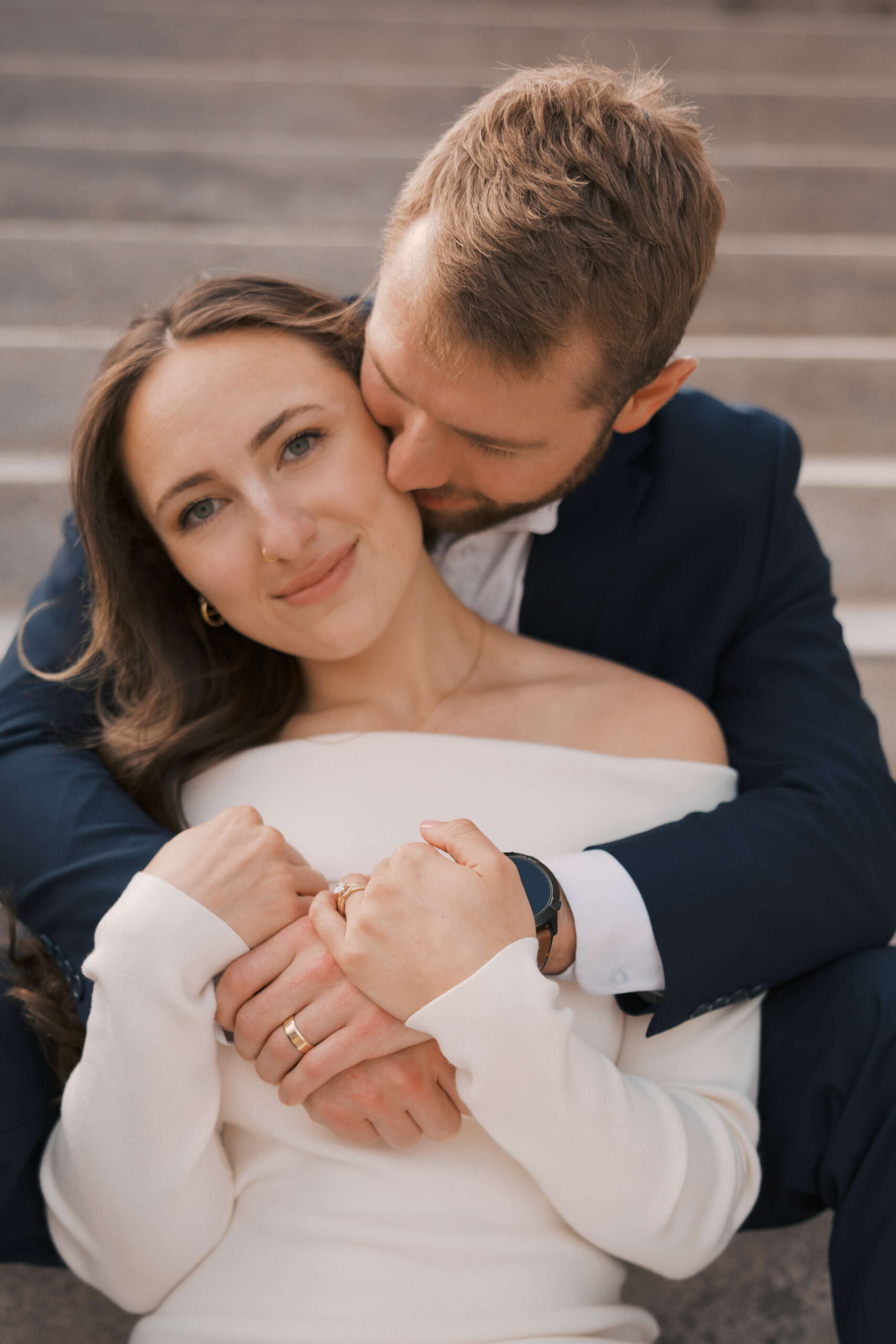 A bride smiles while snuggling in the lap of her groom as he kisses her cheek at their joslyn art museum wedding
