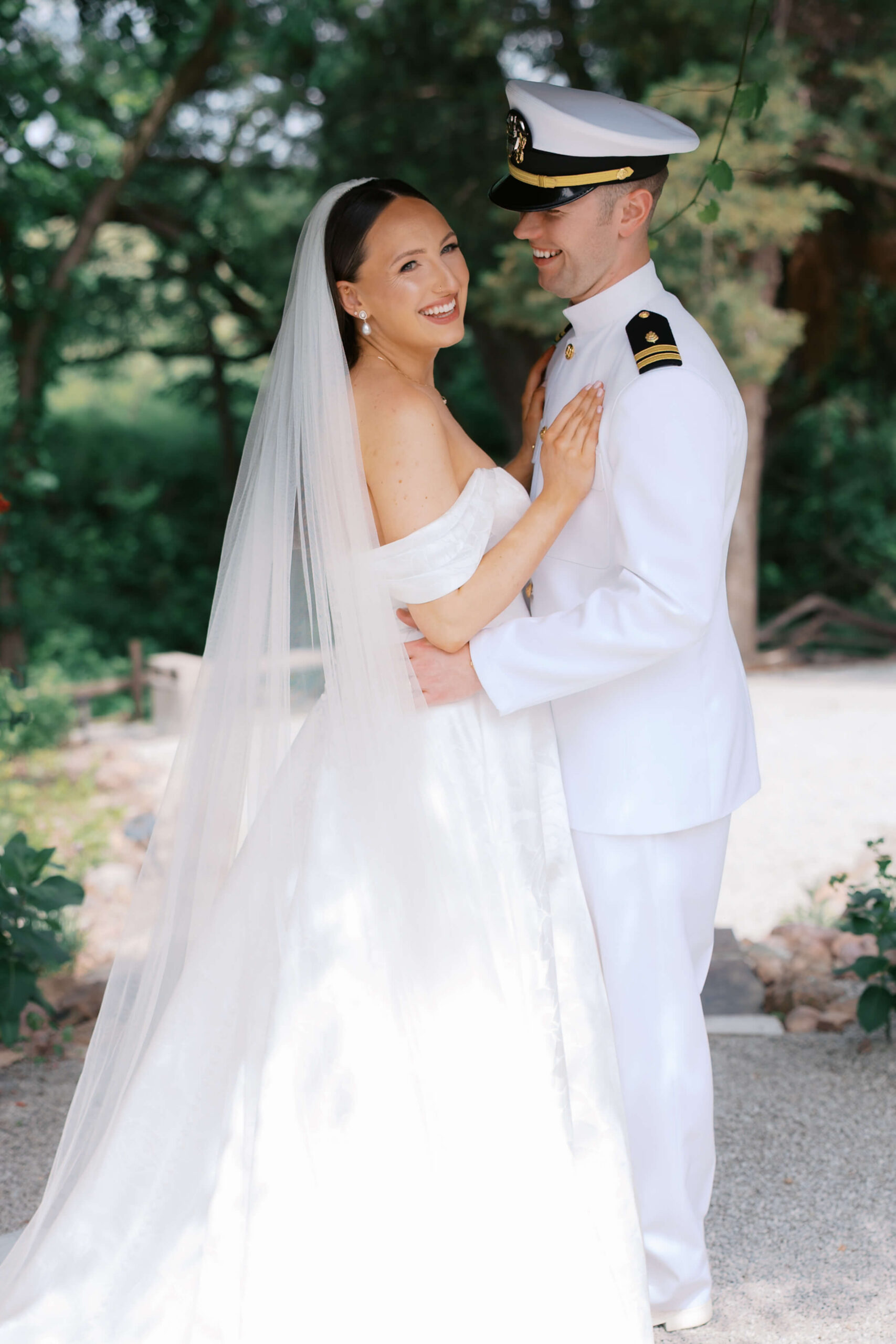 A bride laughs while cuddling with her happy military husband under trees on a patio