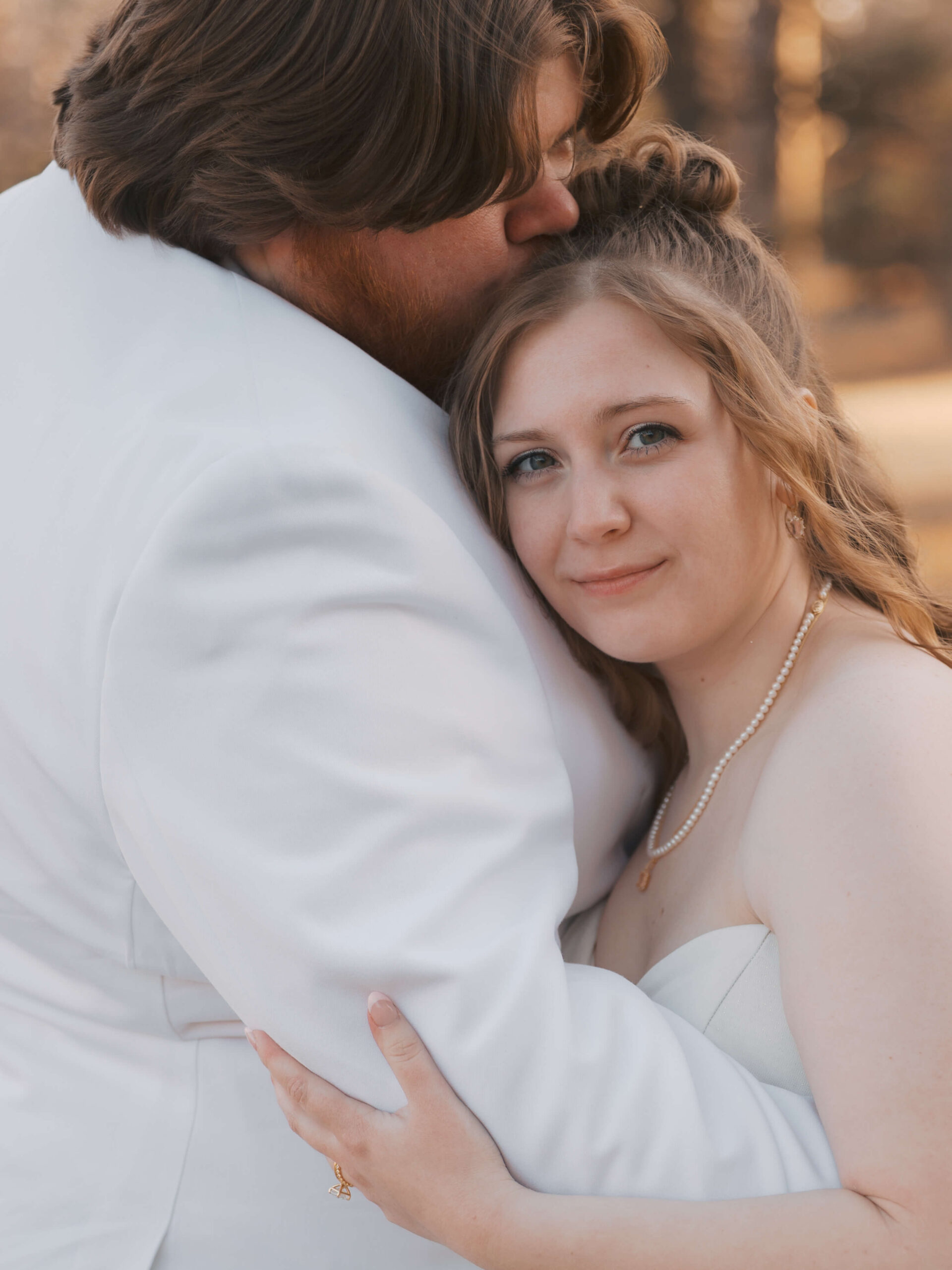 A happy bride leans into her groom's white tux jacket as he kisses her head at sunset