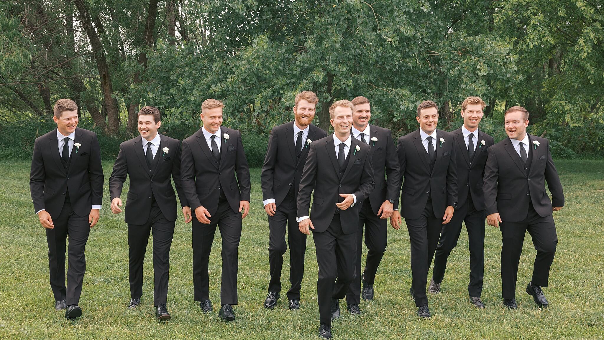 A groom walks with his groomsmen in all matching black tuxedos in a lawn while laughing