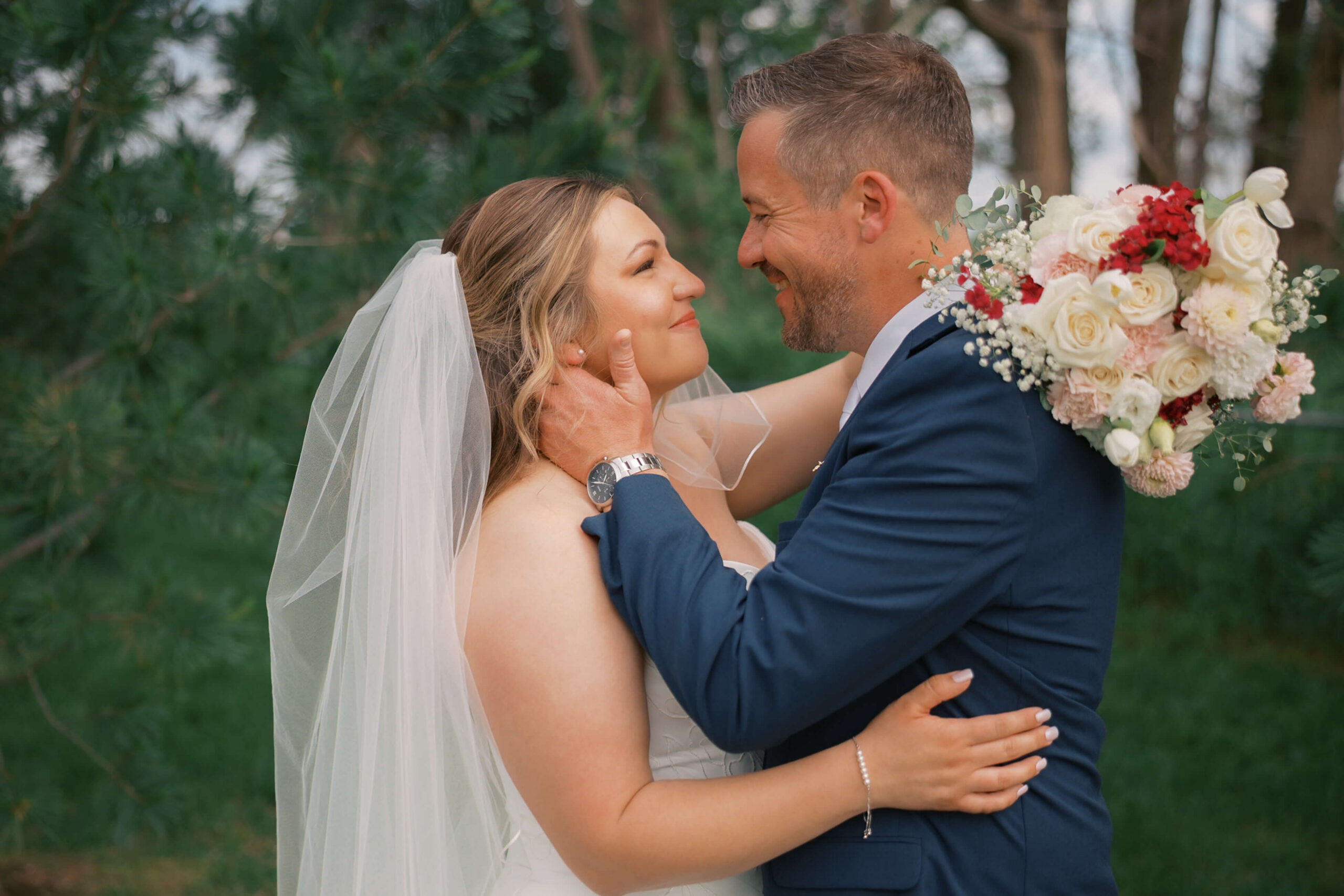 Happy newlyweds smile at each other with the bouquet behind the groom while leaning in for a kiss in a blue suit during his bosana wedding
