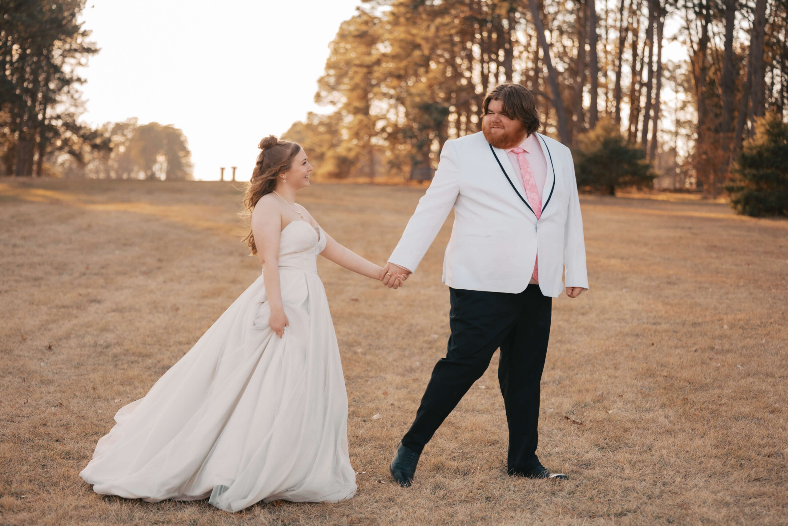 A groom with a white jacket and pink tie leads his bride by the hand through a meadow at sunset at one of the lincoln wedding venues