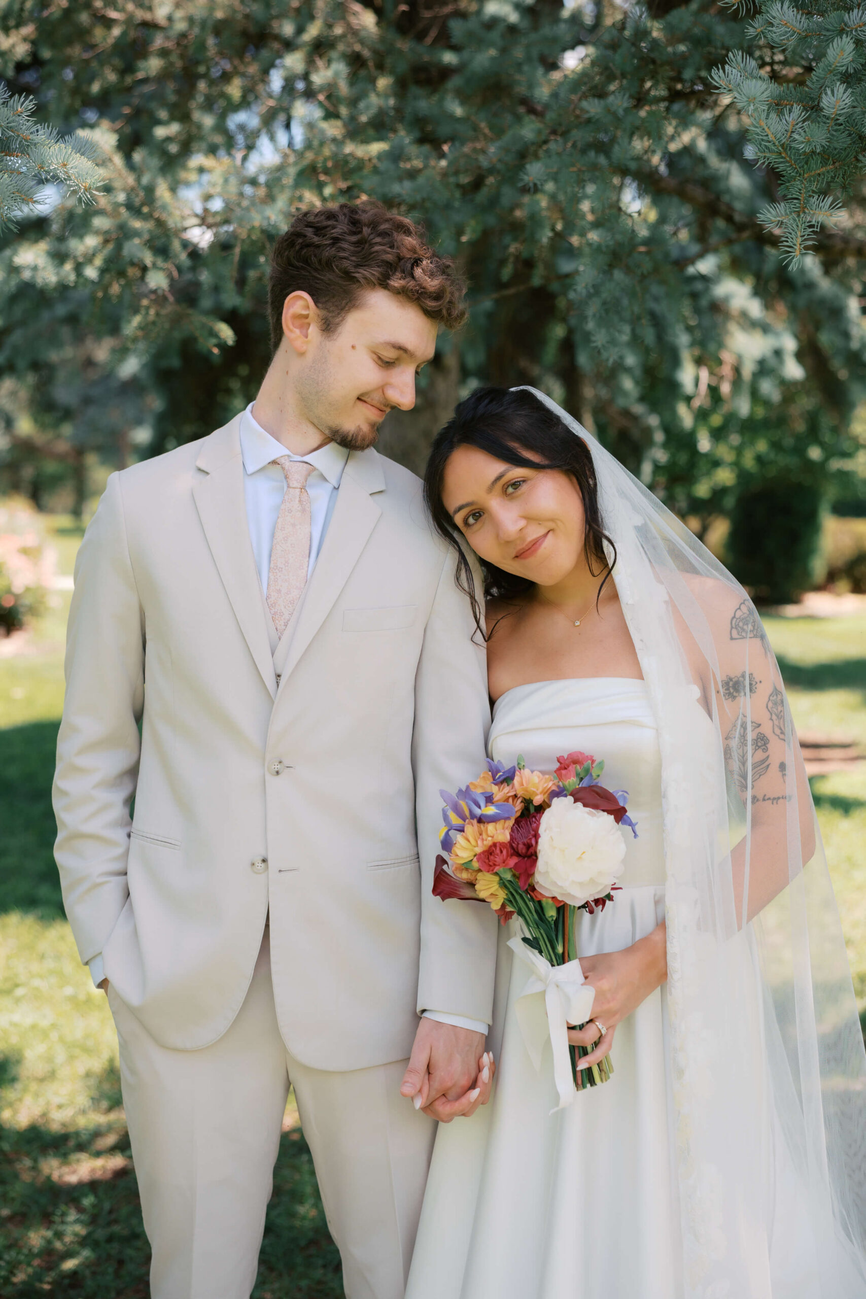 A bride leans on the shoulder of her groom in tan as they stand under pine trees