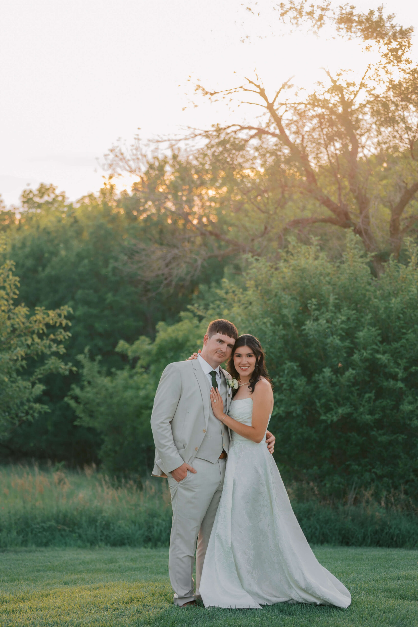 A bride and groom snuggle while standing in a lawn at sunset