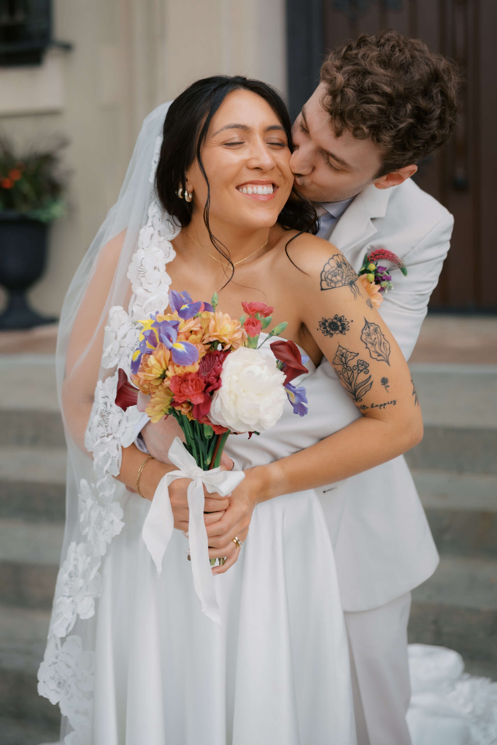 A bride smiles big as her groom in grey hugs and kisses her cheek from behind during their sheldon museum wedding