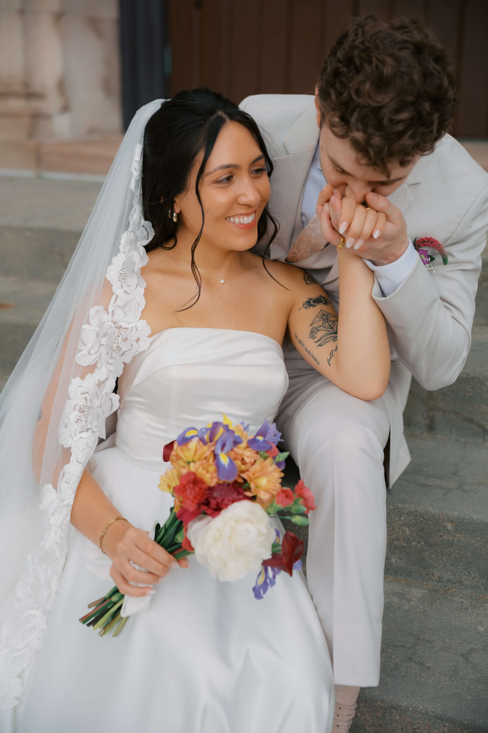 A bride and groom snuggle on stone steps while groom kisses her hand during their sheldon museum wedding