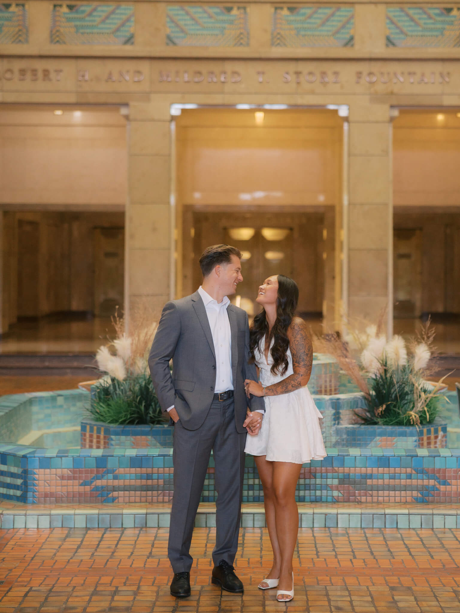 An engaged couple laugh and lean in for a kiss by an atrium fountain