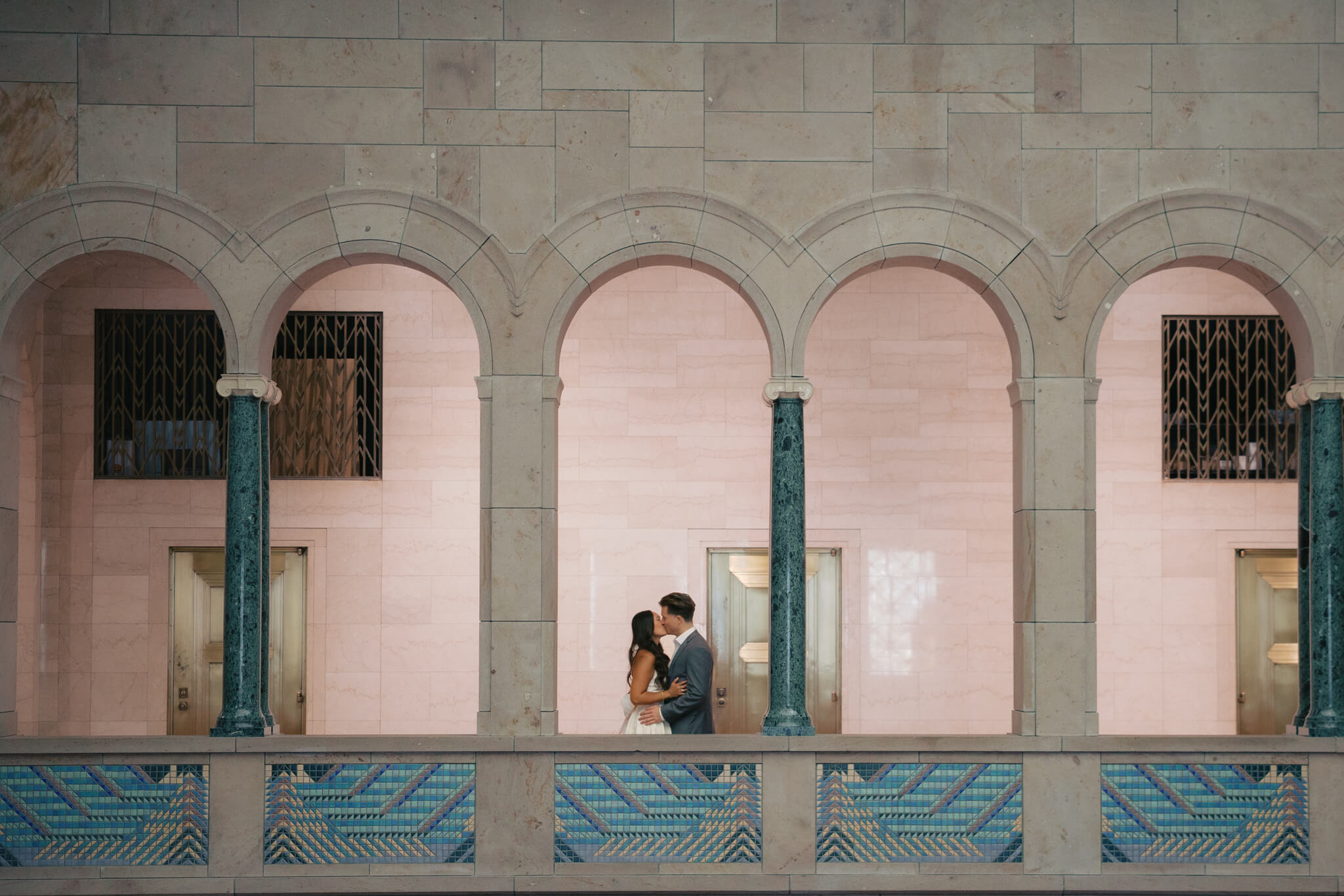 A newly engaged couple kisses under an arch on a museum balcony