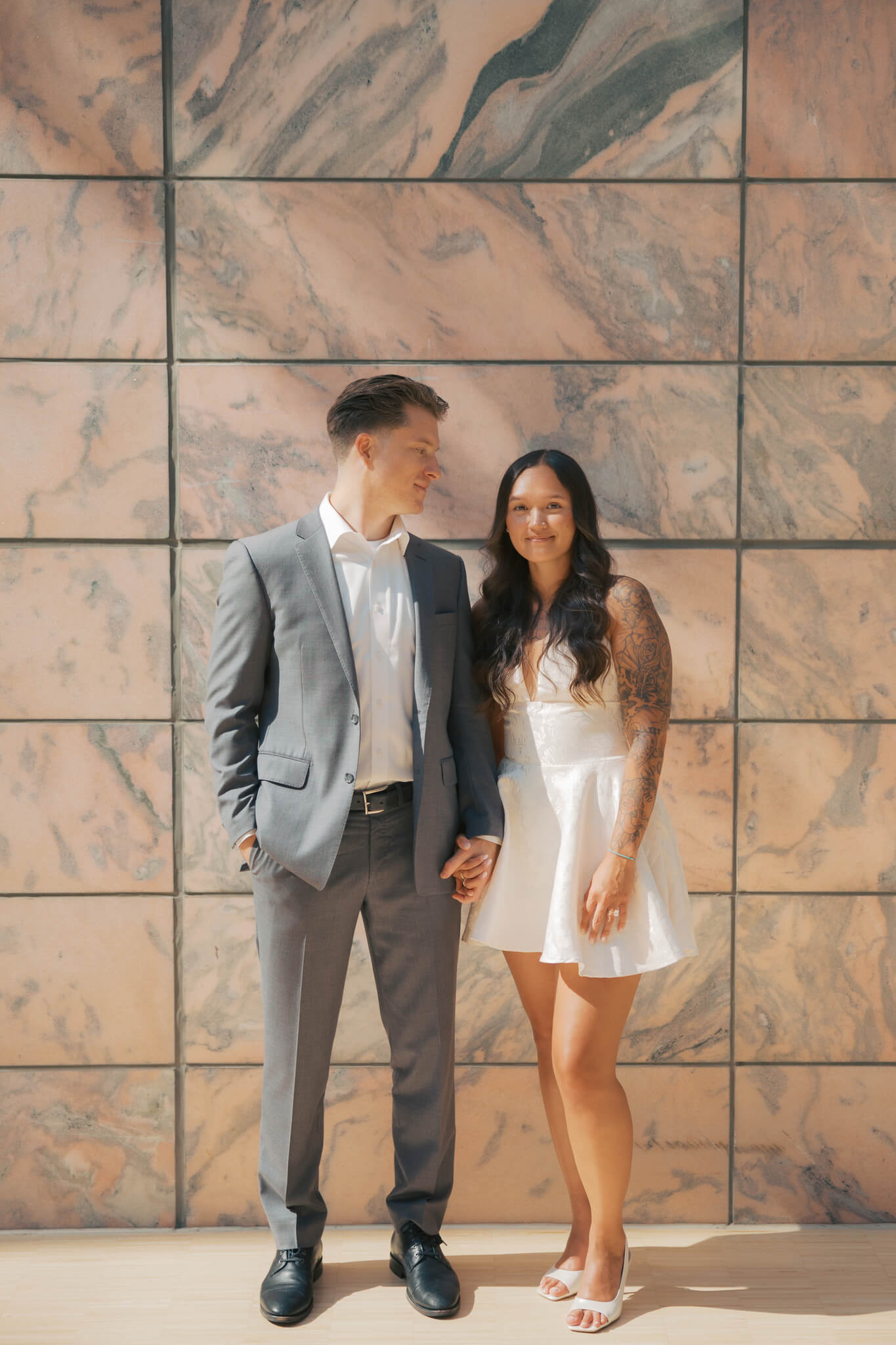 A happy man in a grey suit smiles to his happy fiancee in a white dress as they stand against marble wall