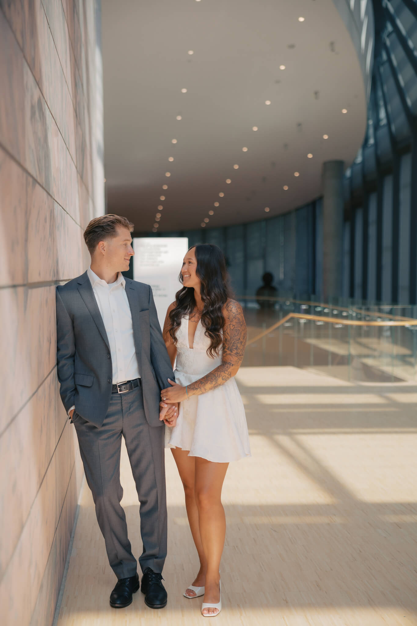 A happy couple lean on the marble walls of the joslyn art museum wedding venue lobby and hold hands smiling at each other