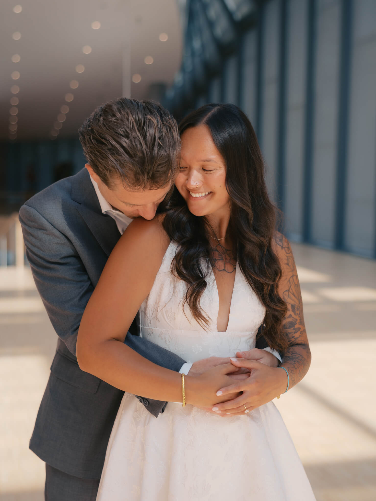 A man in a grey suit kisses the shoulder of his bride as she giggles in a white dress at the joslyn art museum wedding venue
